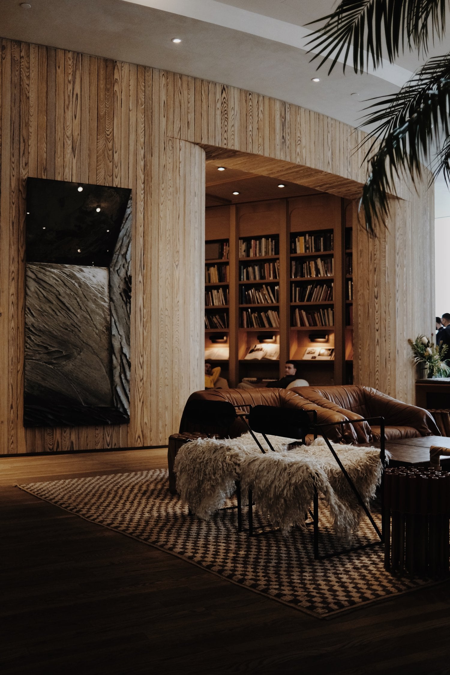 Santa Monica Proper, North America — Wooden-paneled lounge with leather sofa, sheepskin chair, built-in library nook, and black artwork under warm recessed lighting.