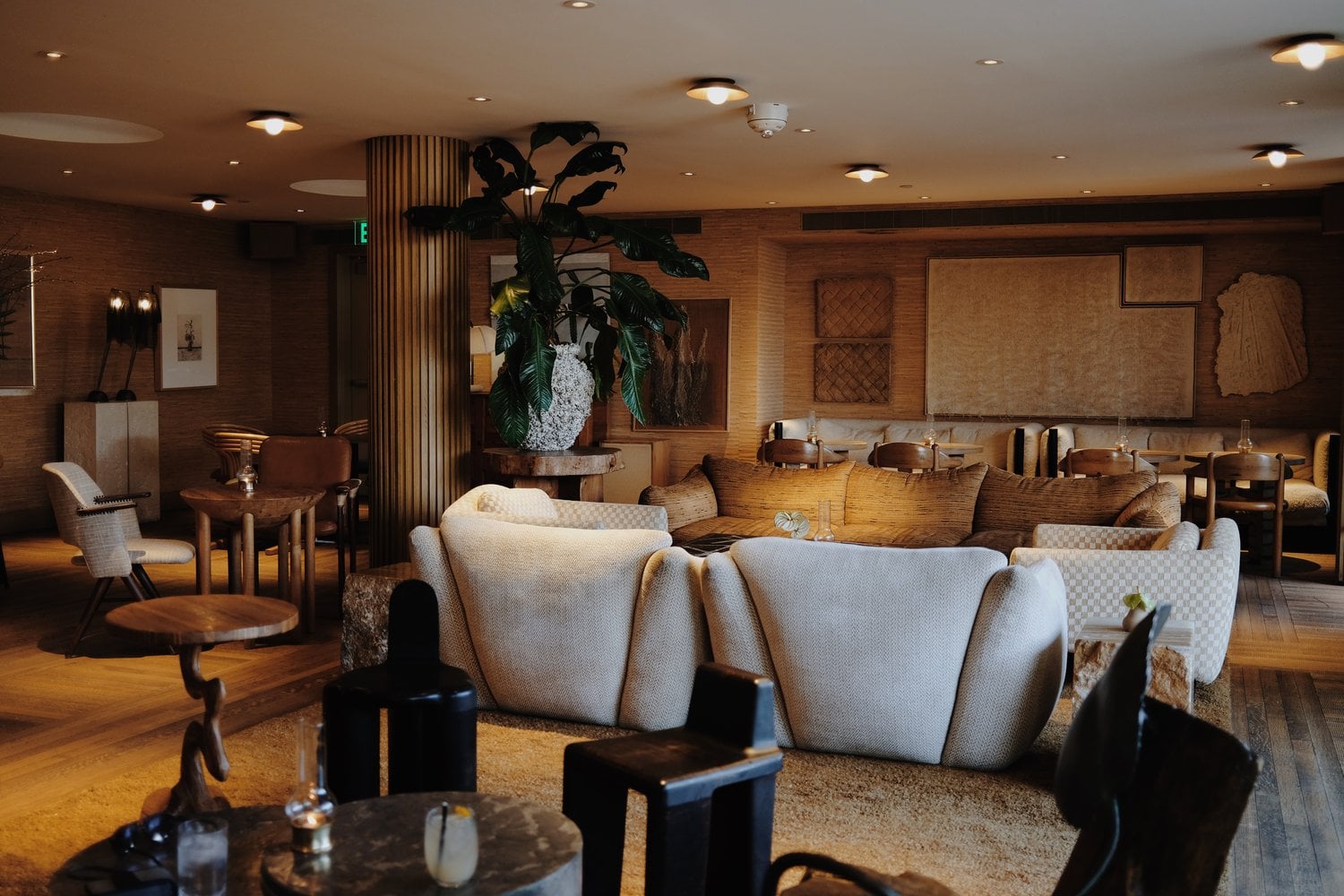 Santa Monica Proper, North America — Warm lounge with cream upholstered seating, exposed brick walls, wooden flooring, brass accents, and potted plants under recessed lighting.