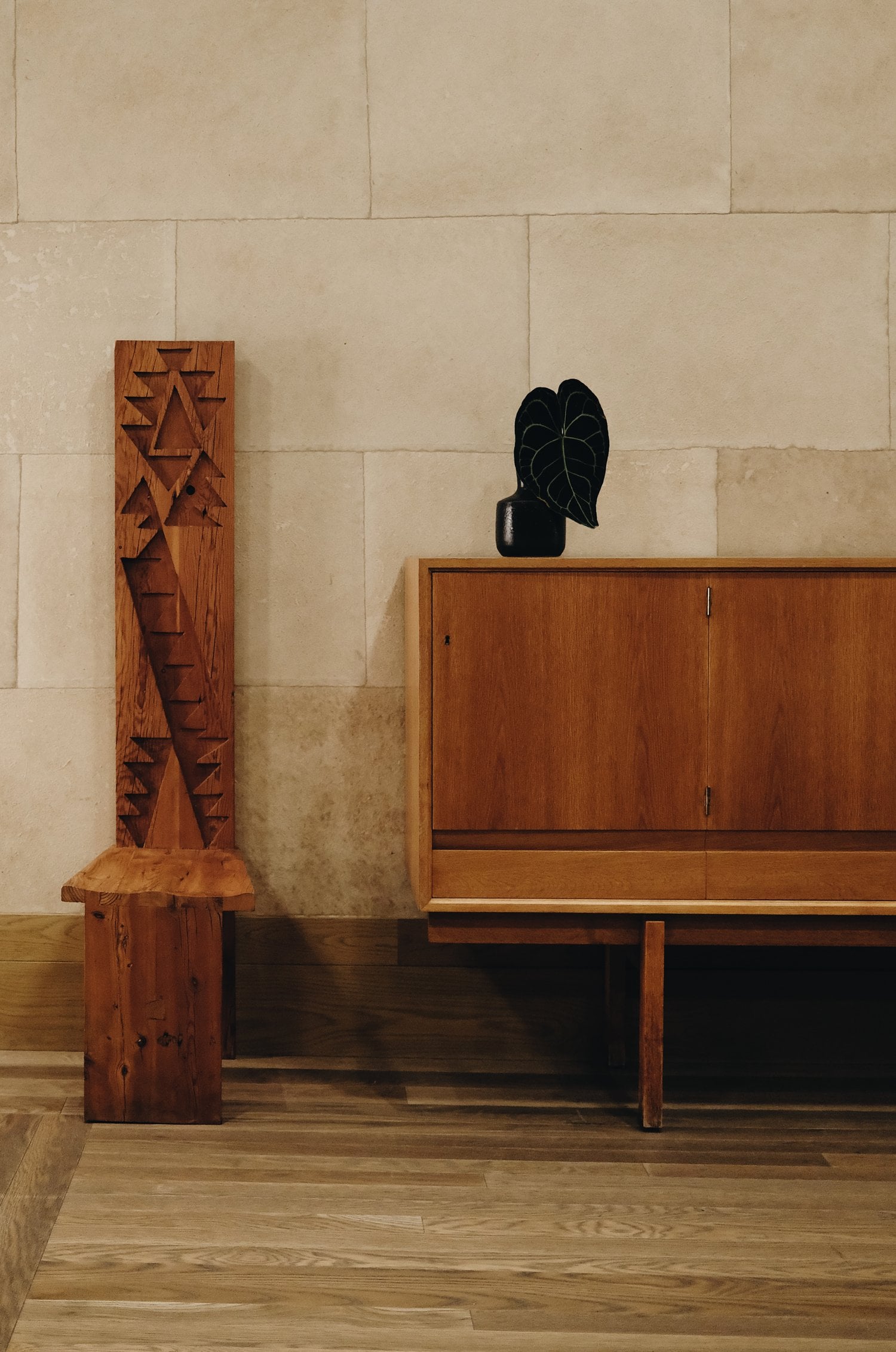 Santa Monica Proper, North America — Wooden sculptural panel and mid-century cabinet with black potted plant against stone wall, warm gallery lighting.