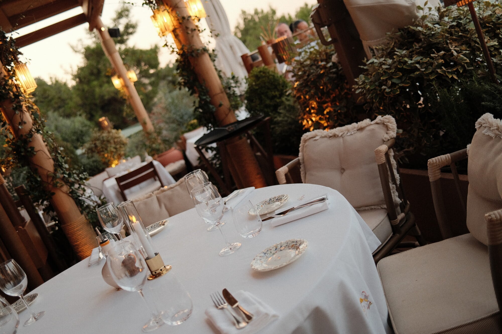 Gigi Bodrum, Asia — Candlelit outdoor dining terrace with round table, cream upholstered chairs, ivy-draped pergola, golden pendant lights, and lush greenery at dusk.