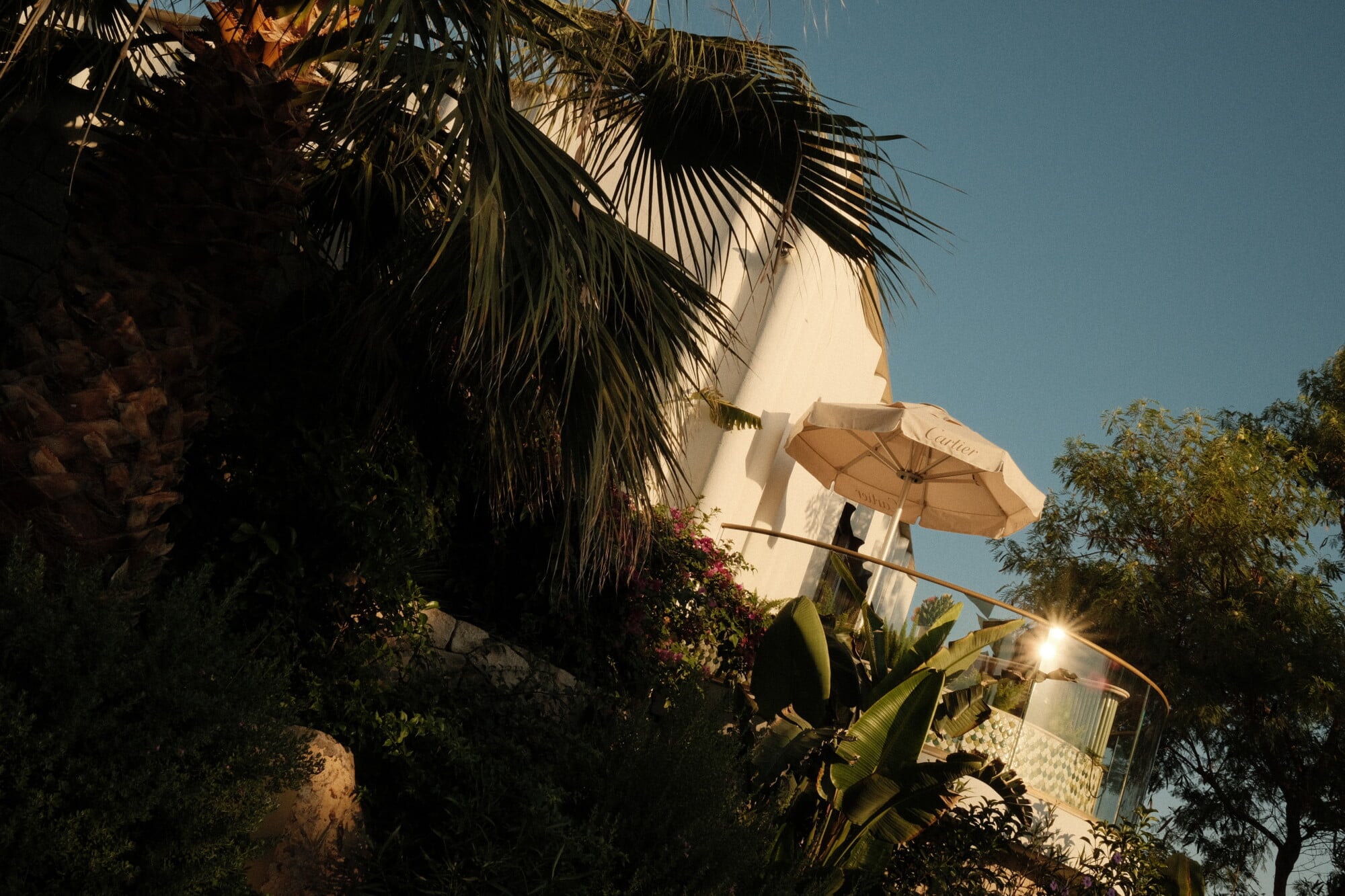 Cartier, Asia — Luxury whitewashed villa with palm fronds, beige umbrella, and flowering vines under clear blue sky, golden hour light.