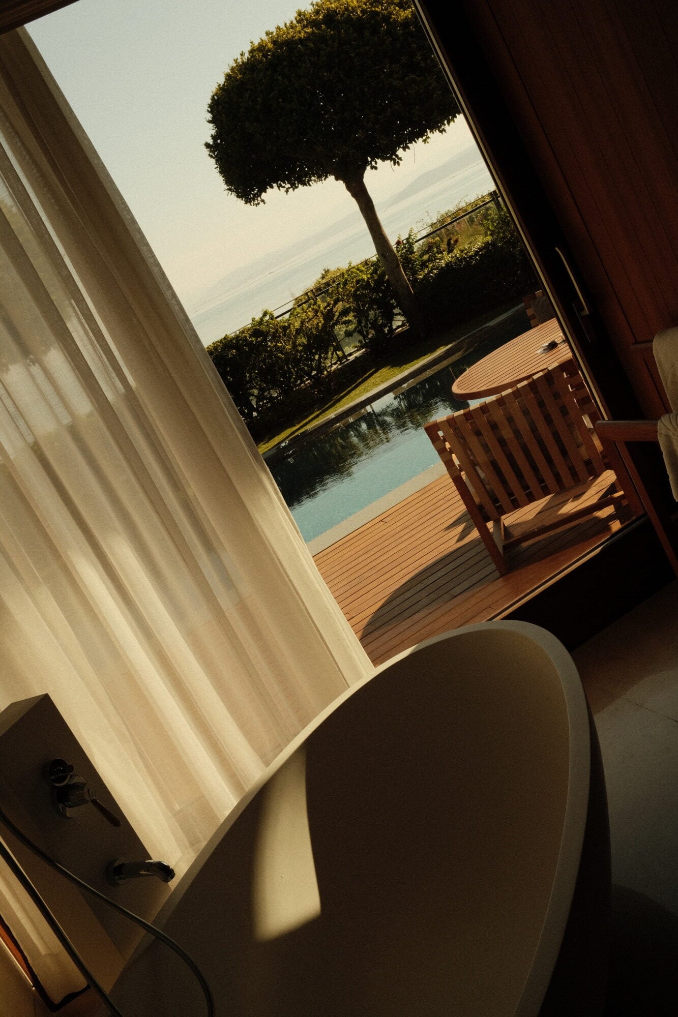 Mandarin Oriental Bodrum, Turkey — Sunlit bedroom with sheer curtains framing terrace view of manicured gardens, pool, and calm water beyond.