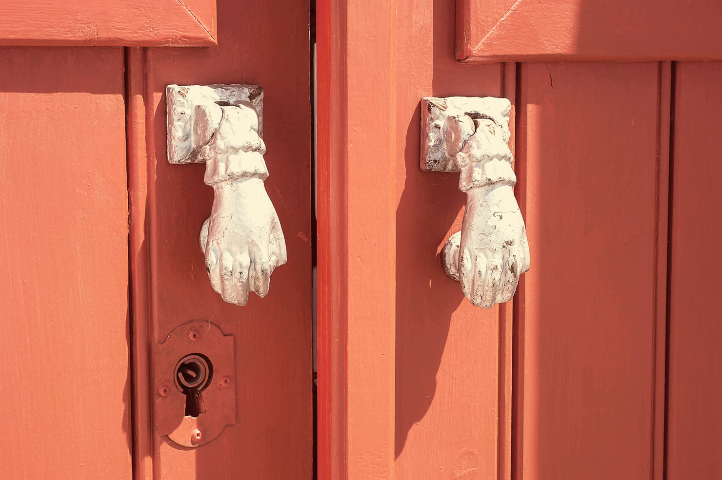 Pensão Agrícola, Europe — Weathered white hand-shaped door knockers on coral-red painted wooden doors with ornate keyhole detail.