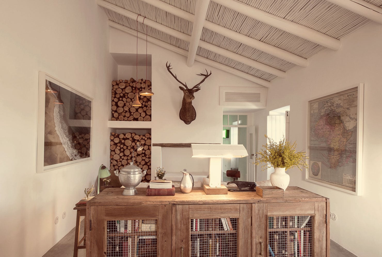Pensão Agrícola, Europe — Bright office with exposed beam ceiling, wood desk, taxidermy deer head, vintage maps, stacked wood wall art, and natural light from skylights.