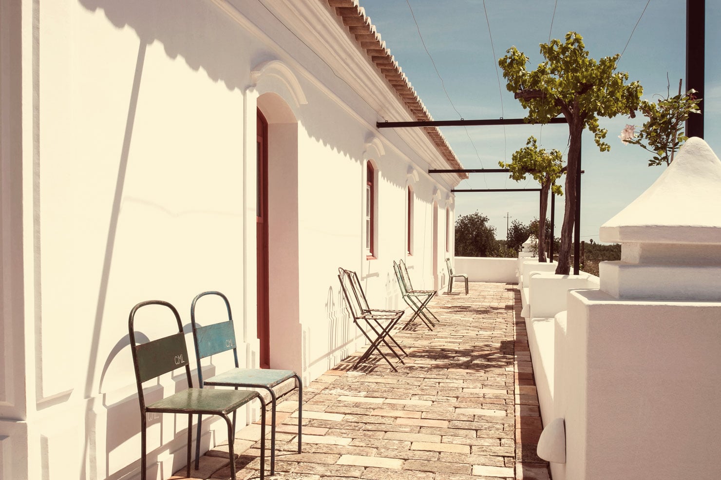 Pensão Agrícola, Europe — Whitewashed terrace with wooden pergola, metal chairs, terracotta roof, Mediterranean coastal architecture and shadows at golden hour.