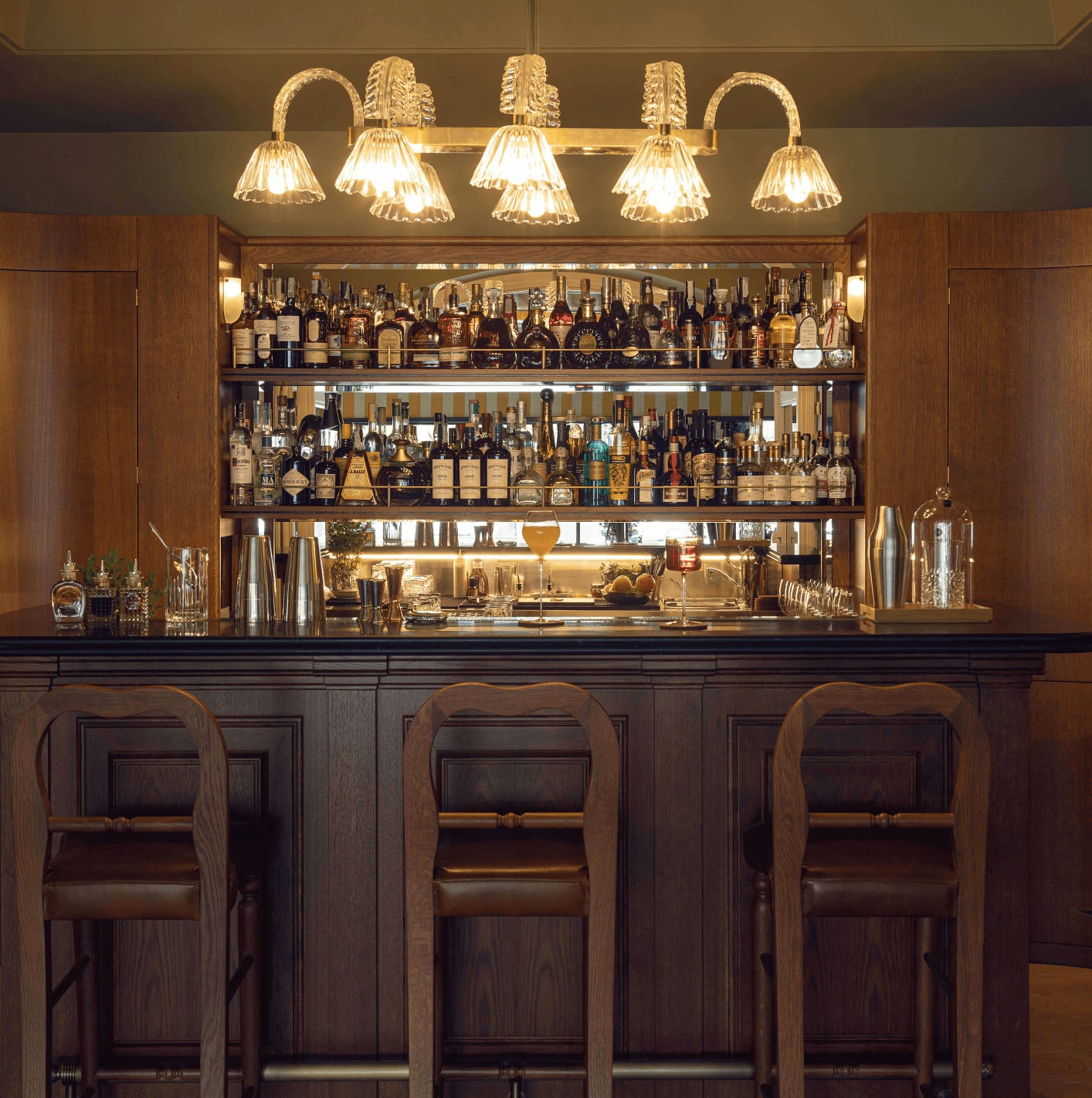 DaV Mare Bar, Europe — Illuminated back bar with brass five-light fixture, walnut cabinetry, three bar stools, and extensive spirit collection displayed across mirrored shelving.