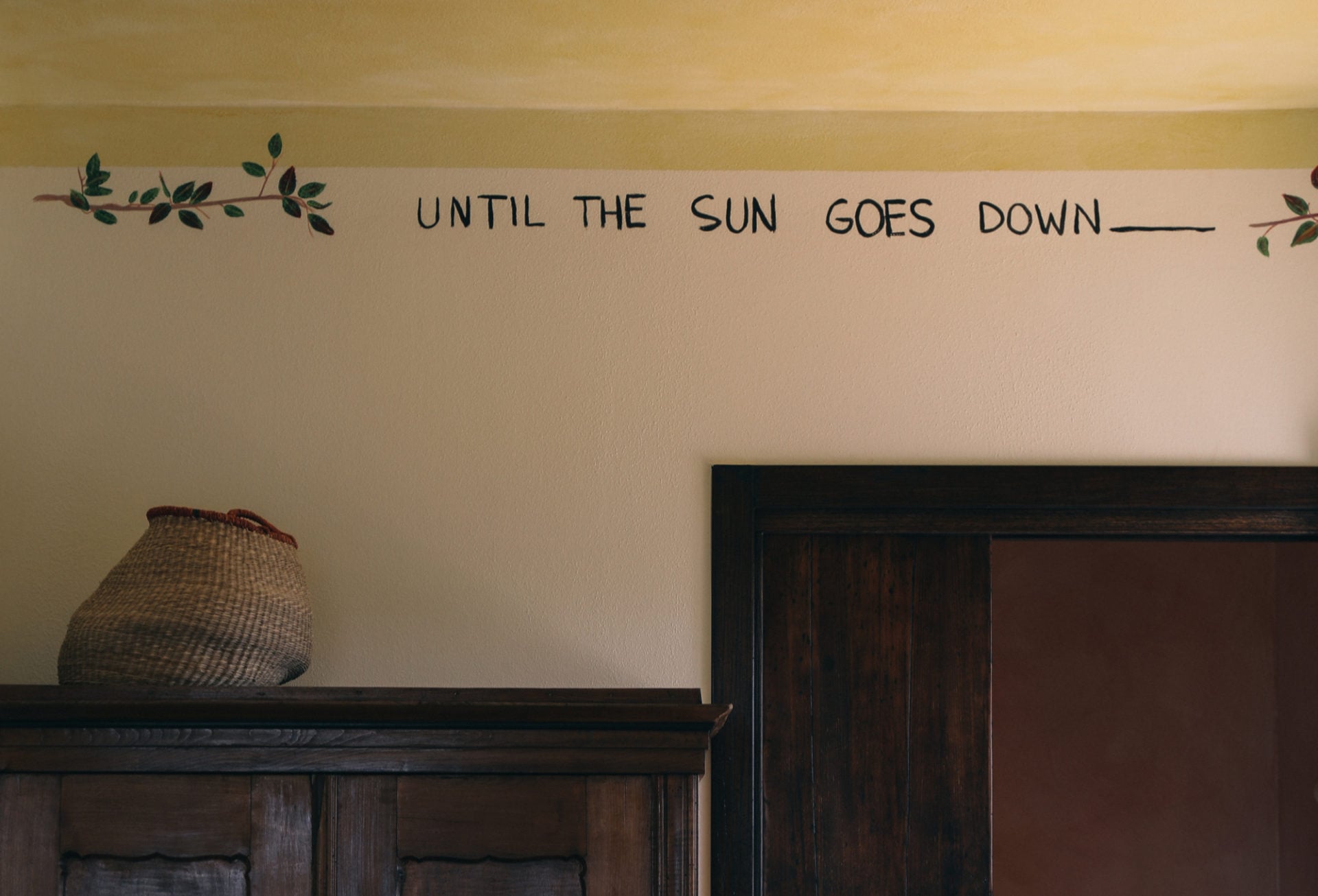 Villa Lena, Europe — Wall-mounted typography with leaf motifs above woven basket on dark wooden shelf, minimalist hospitality interior design.
