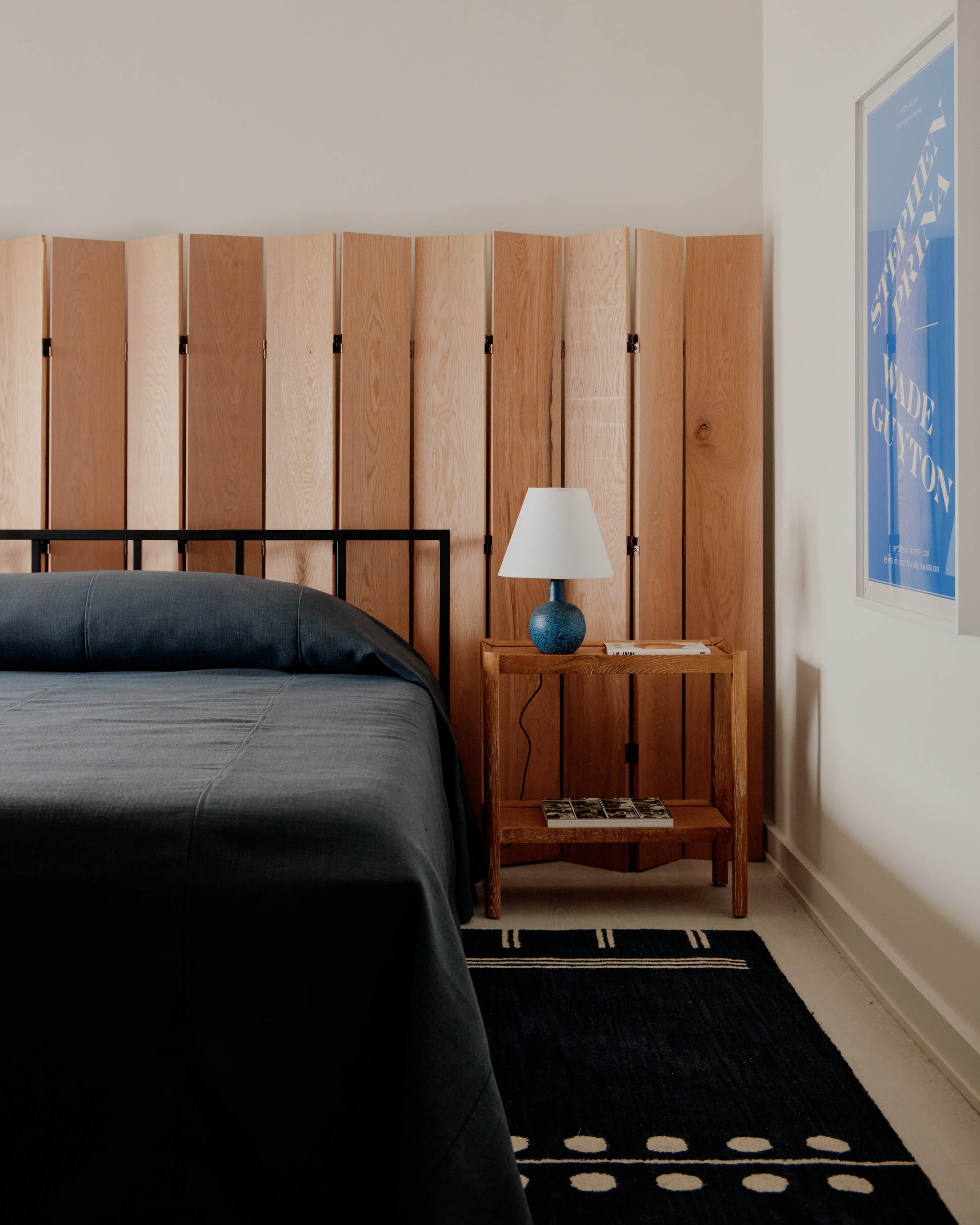 The James Bradley, North America — Minimalist bedroom with black metal bed frame, charcoal linens, wooden room divider screen, teak nightstand with blue ceramic lamp, geometric black rug, white walls, and blue artwork.