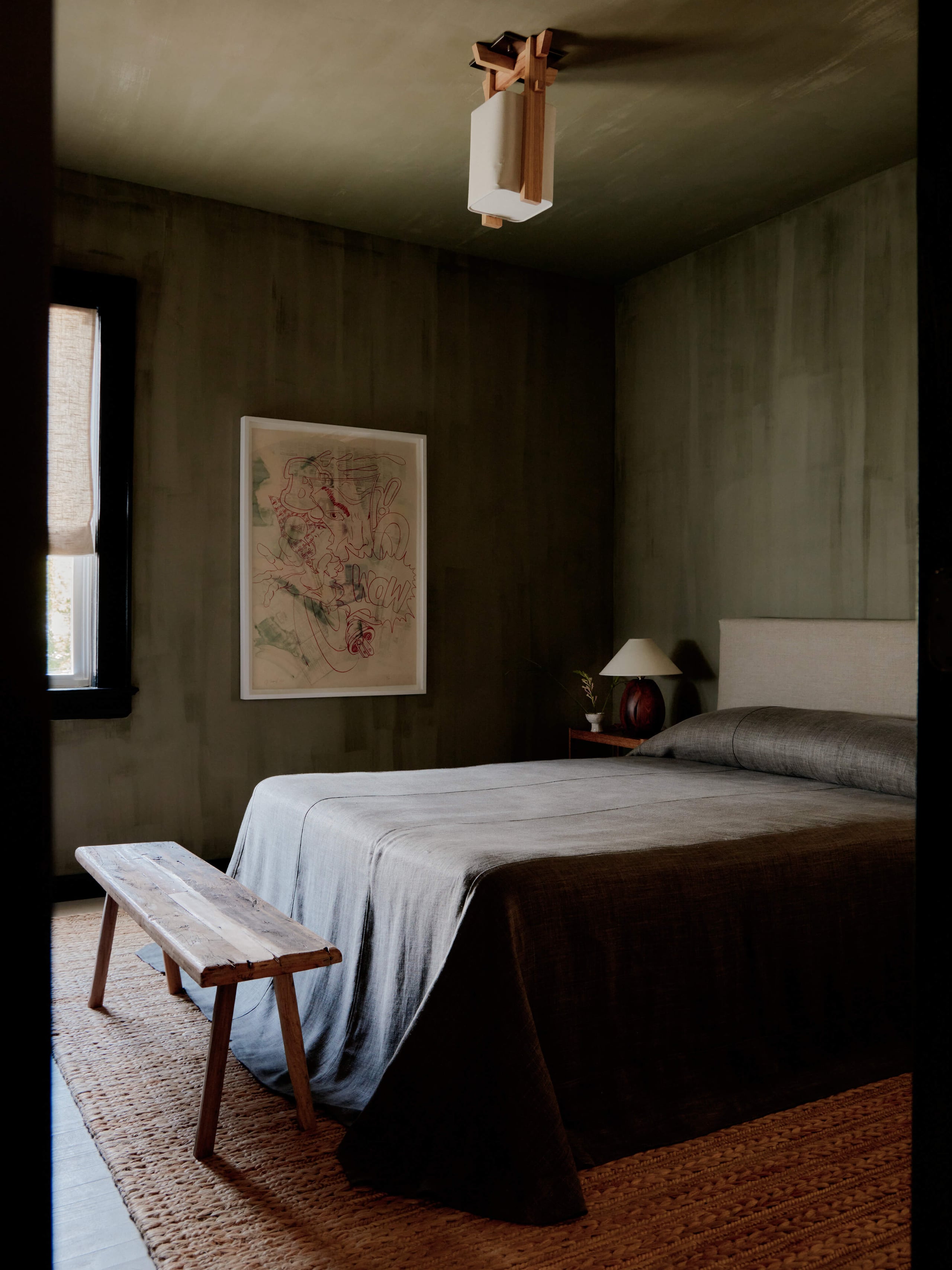 The James Bradley, North America — Minimalist hotel bedroom with olive walls, wooden bed frame, linen bedding, mid-century bench, framed artwork, and warm natural light from narrow window.