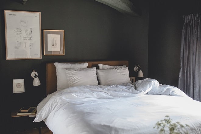 Le Barn, Europe — Minimalist bedroom with charcoal walls, wooden headboard, white linens, framed sketches, grey curtains, and soft natural light.