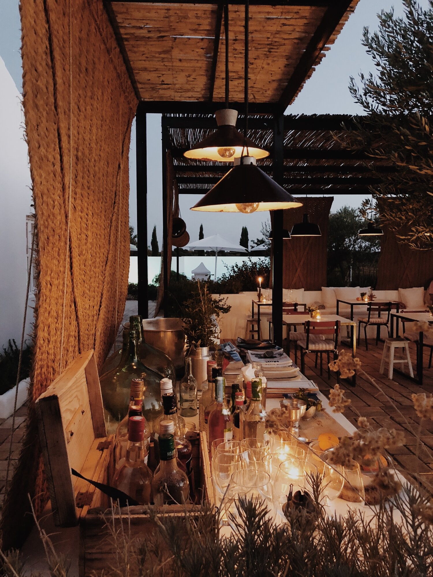 Pensão Agrícola, Europe — Twilight terrace with wooden pergola, pendant lamps, brick wall, bottles and glassware on table, cypress trees and distant landscape.