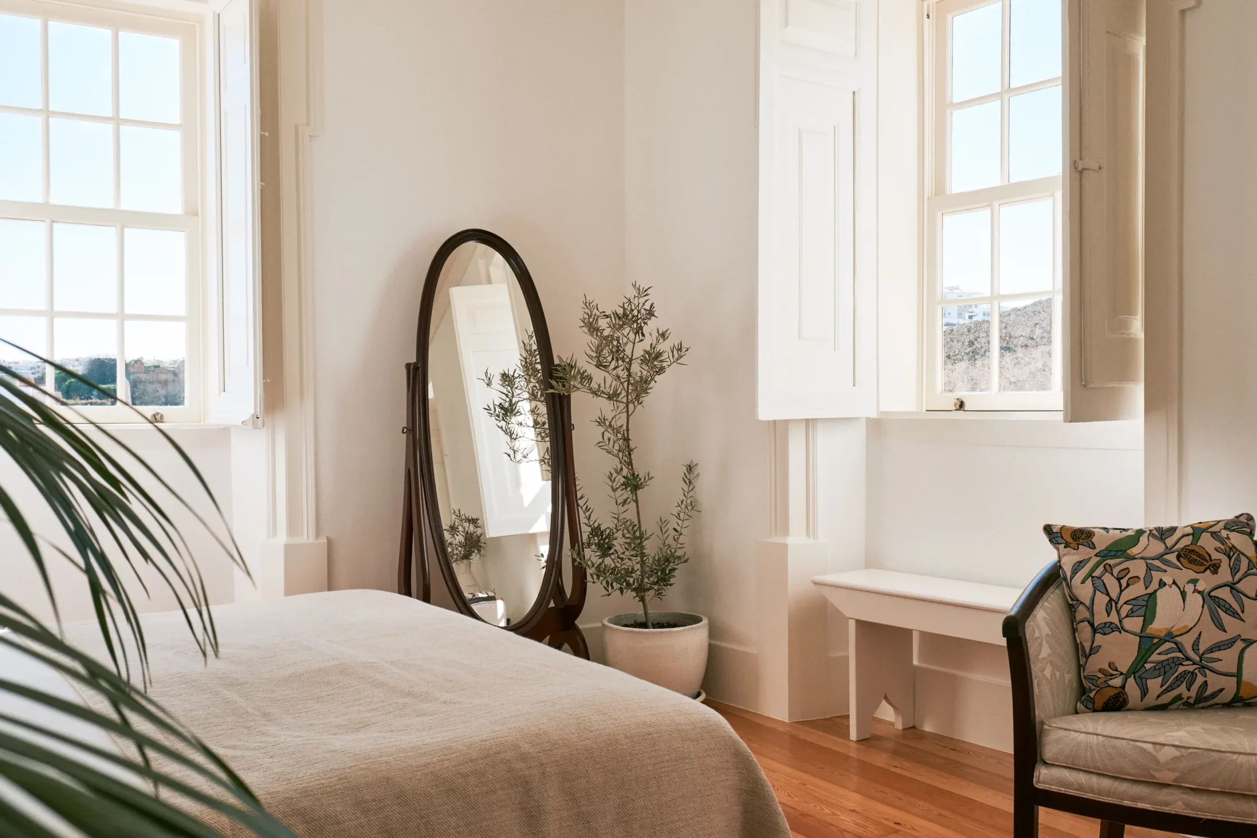 Casa Mãe, Europe — Bright bedroom with arched black mirror, potted plant, wooden flooring, cream bedding, and expansive windows overlooking hillside landscape.