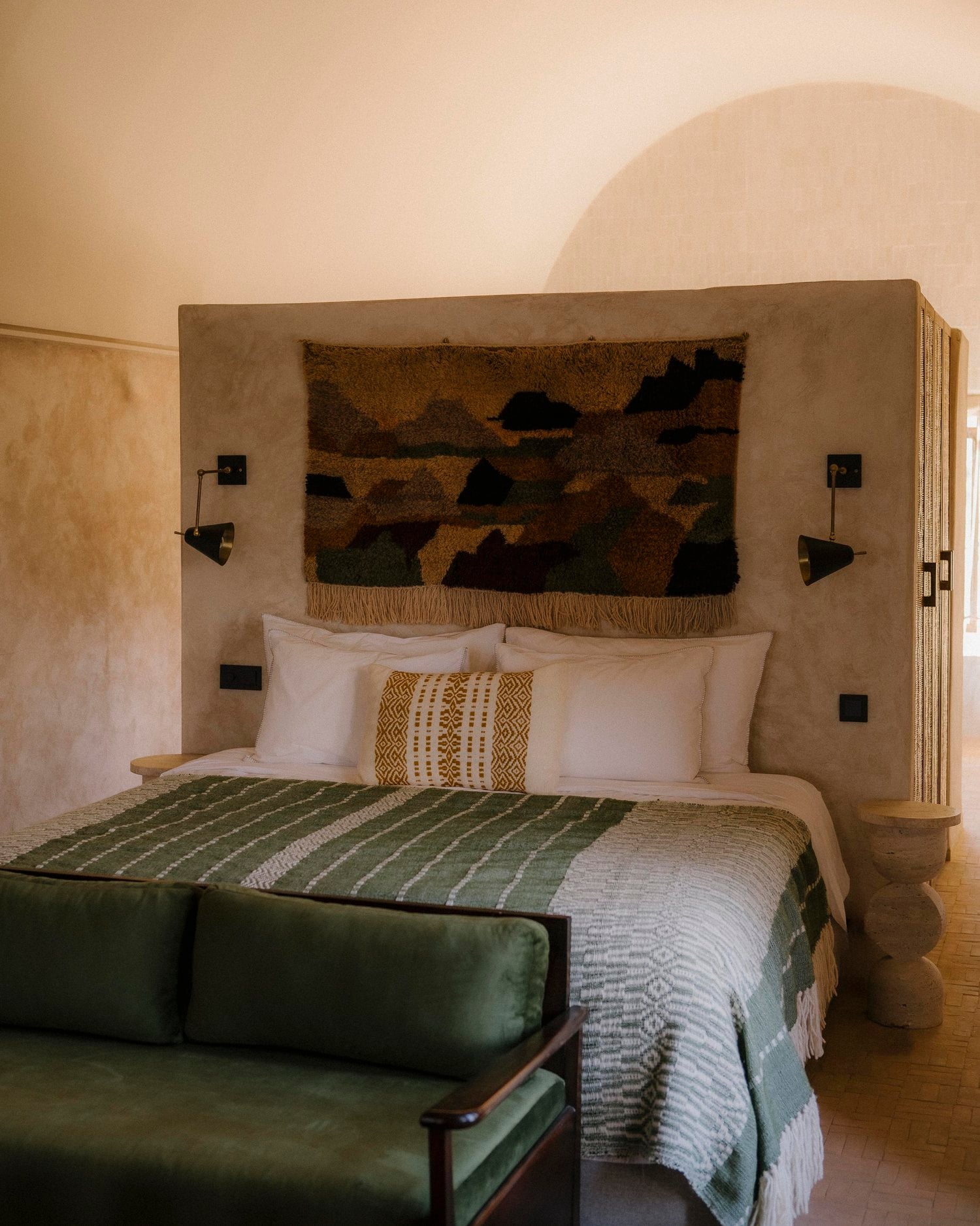 Farasha Farmhouse, Africa — Minimalist bedroom with curved plaster headboard, woven textile art, black task lighting, green striped bedding, and warm earth-tone palette.