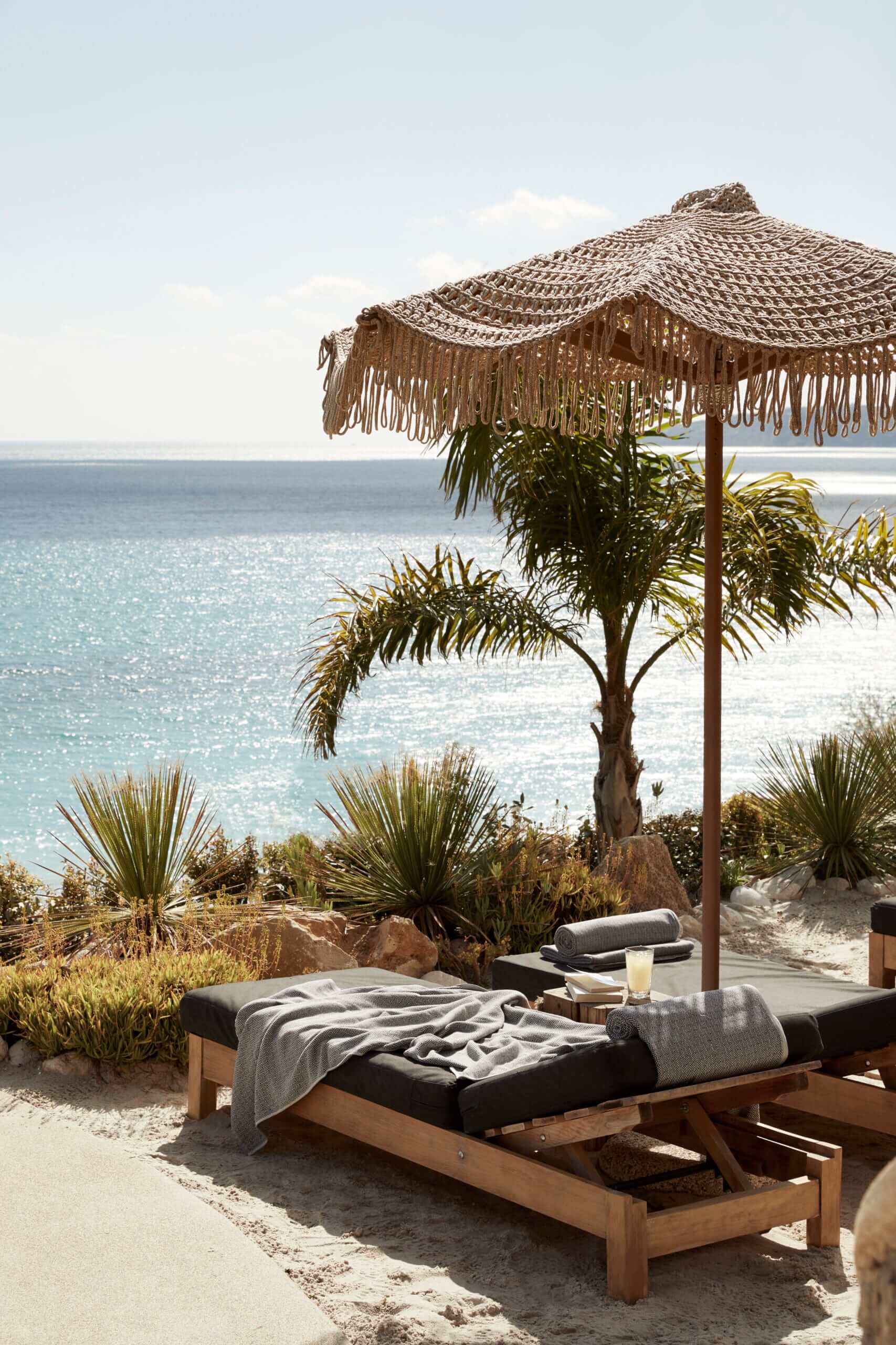 F Zeen Retreat, Europe — Wooden daybed with gray linens beneath a thatched parasol overlooking calm turquoise sea, flanked by palm plants and coastal vegetation.