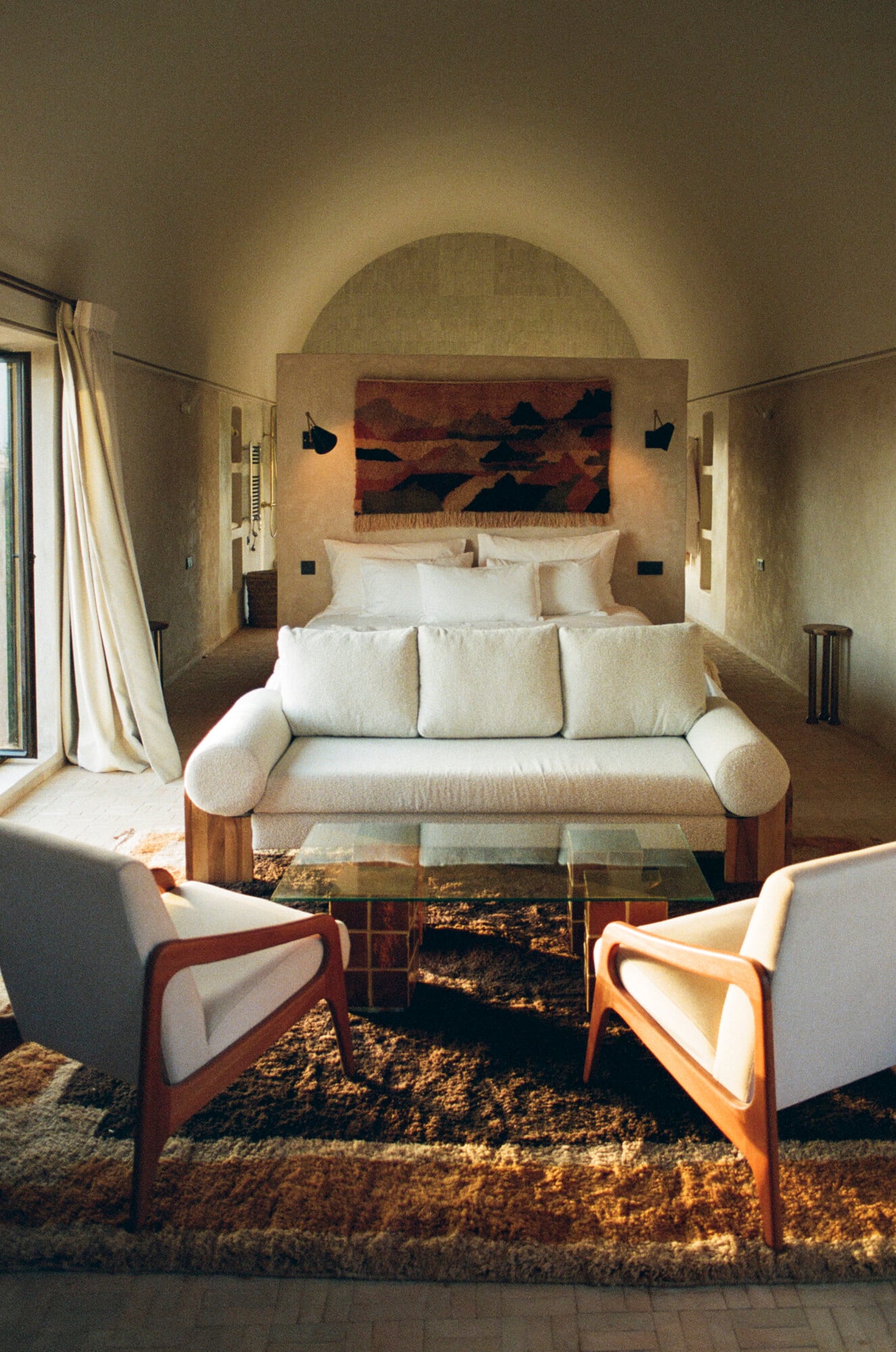 Farasha Farmhouse, Africa — Minimalist bedroom with arched stone walls, cream linen sofa, mid-century wooden armchairs, abstract textile art, warm natural light from large windows.