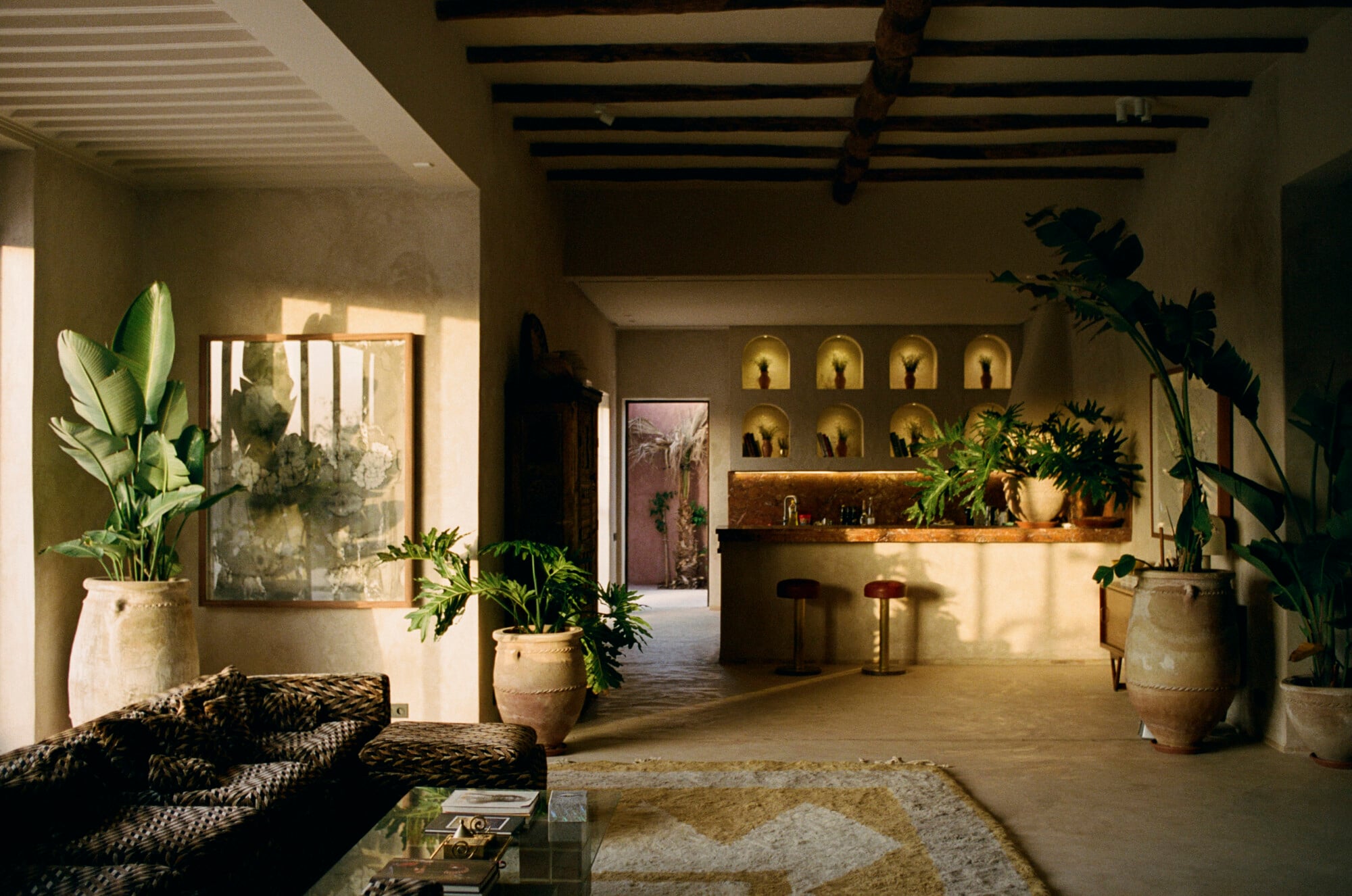 Farasha Farmhouse, Africa — Minimalist interior with earth-toned walls, wooden beams, potted plants, illuminated niches, and natural light casting geometric shadows across terracotta floors.