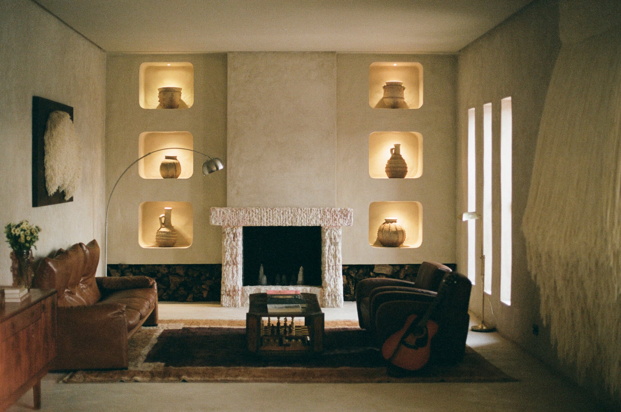 Farasha Farmhouse, Africa — Minimalist living room with illuminated wall niches displaying pottery, textured fireplace surround, leather seating, and brass arc lamp against warm stone walls.