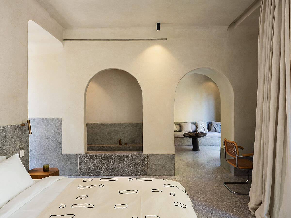 Maison Brummell Majorelle, Africa — Minimalist bedroom with arched concrete alcoves, textured stone wall panels, and natural light creating geometric shadows on cream bedding.