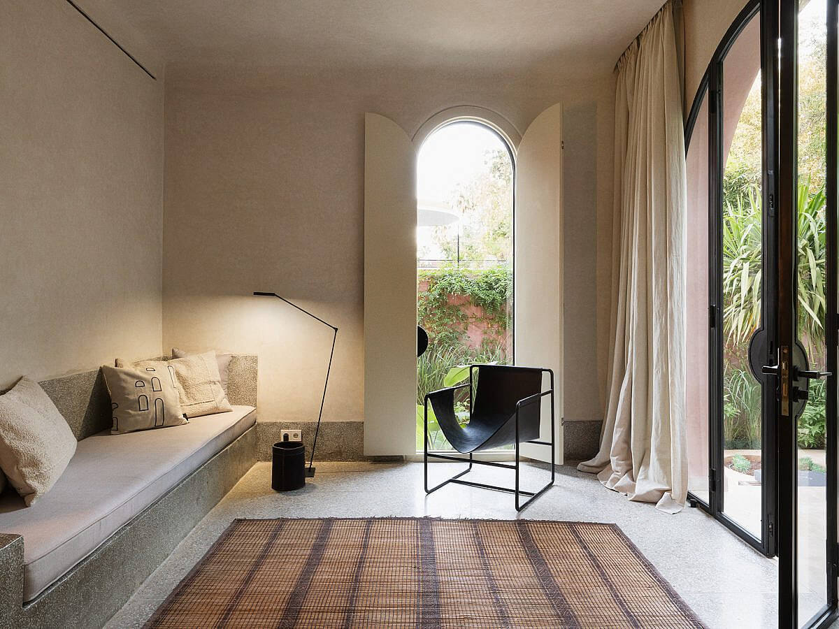 Maison Brummell Majorelle, Africa — Minimalist bedroom alcove with arched window, built-in daybed, black chair, terrazzo flooring, and garden views through glass doors.