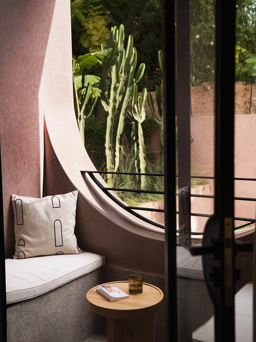 Maison Brummell Majorelle, Africa — Curved terracotta alcove with linen cushions, wooden side table, and view of tall green cactus through black-framed window.