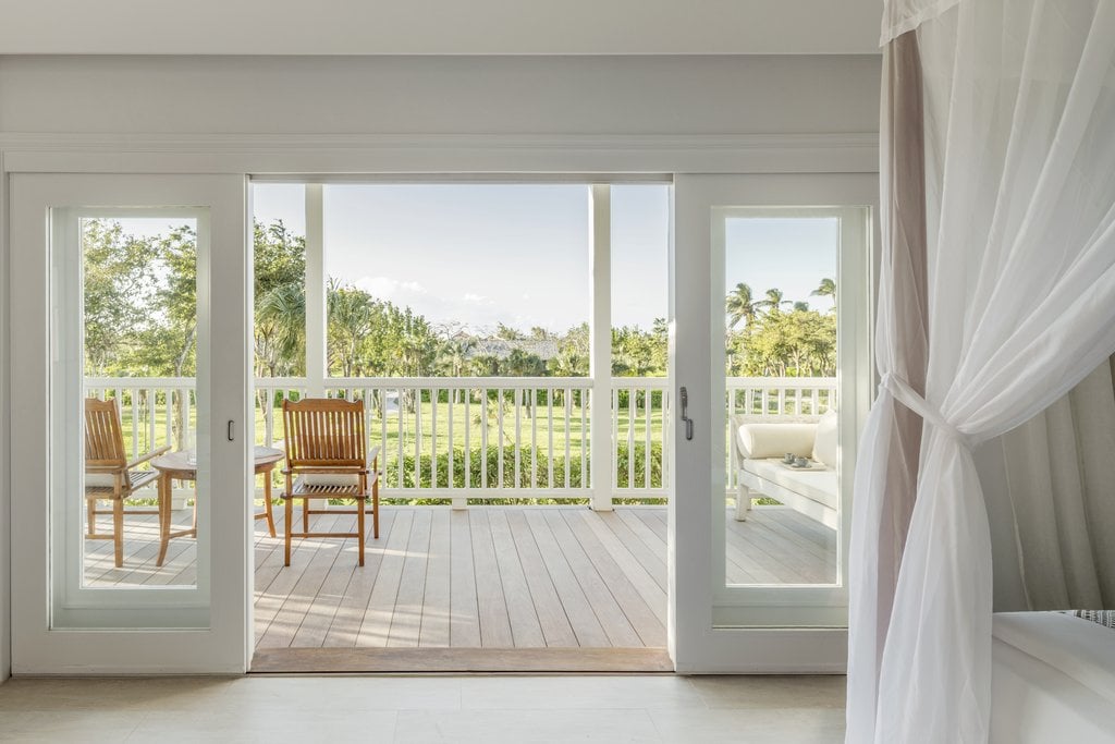 COMO Parrot Cay, North America — Beachfront bedroom with open glass doors revealing wooden deck, rocking chairs, tropical landscape, gauze curtains, and bright natural light.