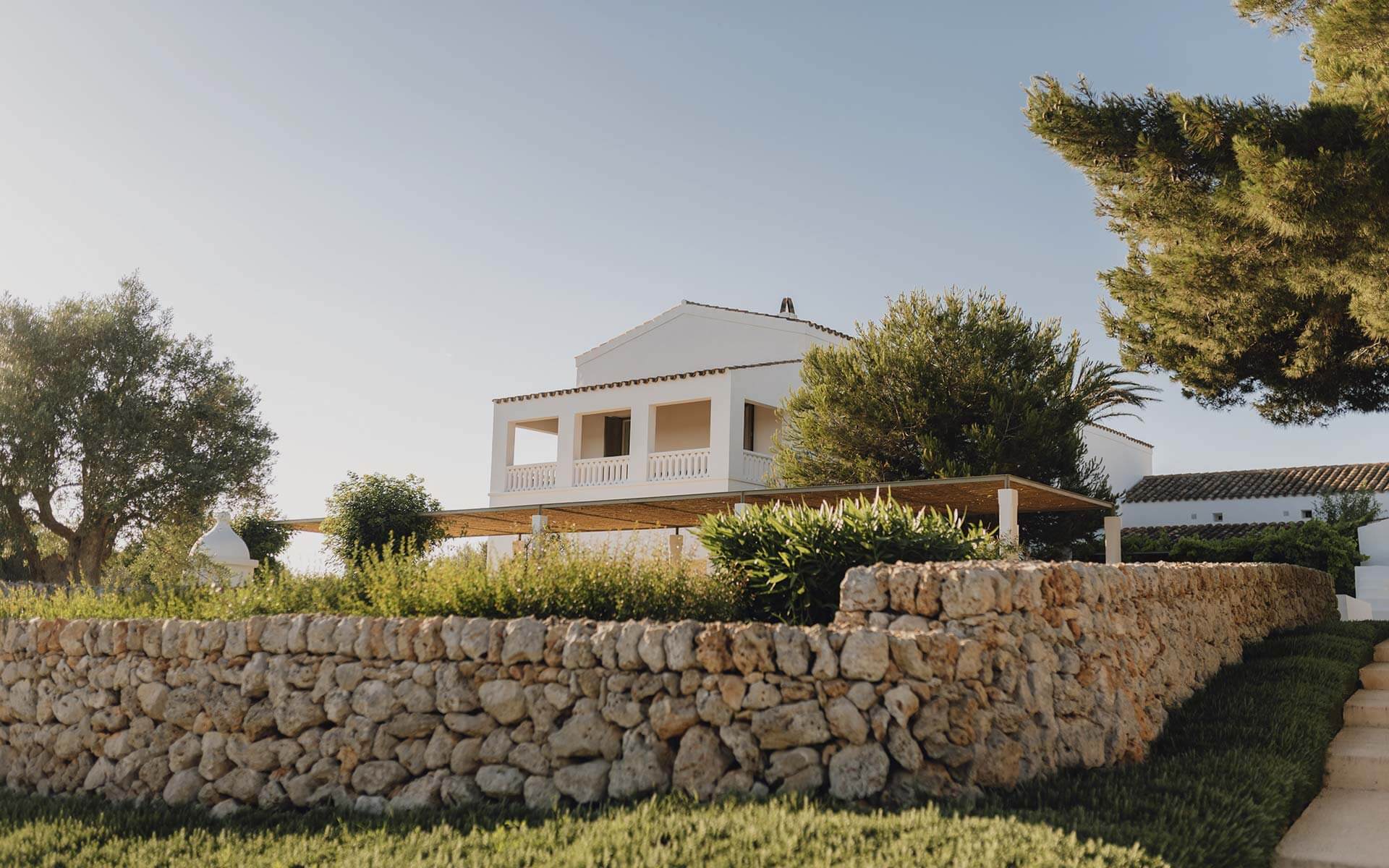 Torralbenc, Balearic Islands, Spain — Whitewashed villa with stone terrace wall, wooden pergola, and Mediterranean gardens under clear sky.