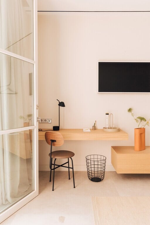 Plácido y Grata, Europe — Minimalist wooden desk workspace with black chair, black monitor, natural light from glass doors, warm beige walls, potted plant, and wire basket.