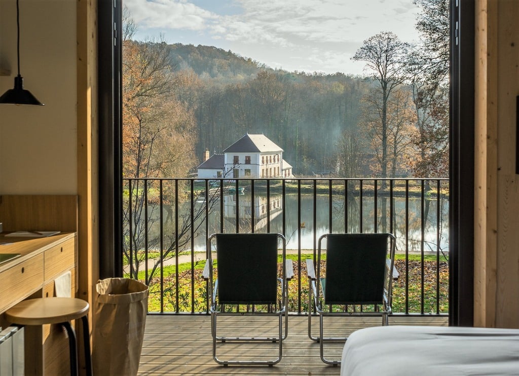 Le Barn, Europe — Modern bedroom with wooden finishes overlooking manicured grounds, river, and historic mansion nestled in forested valley during autumn.