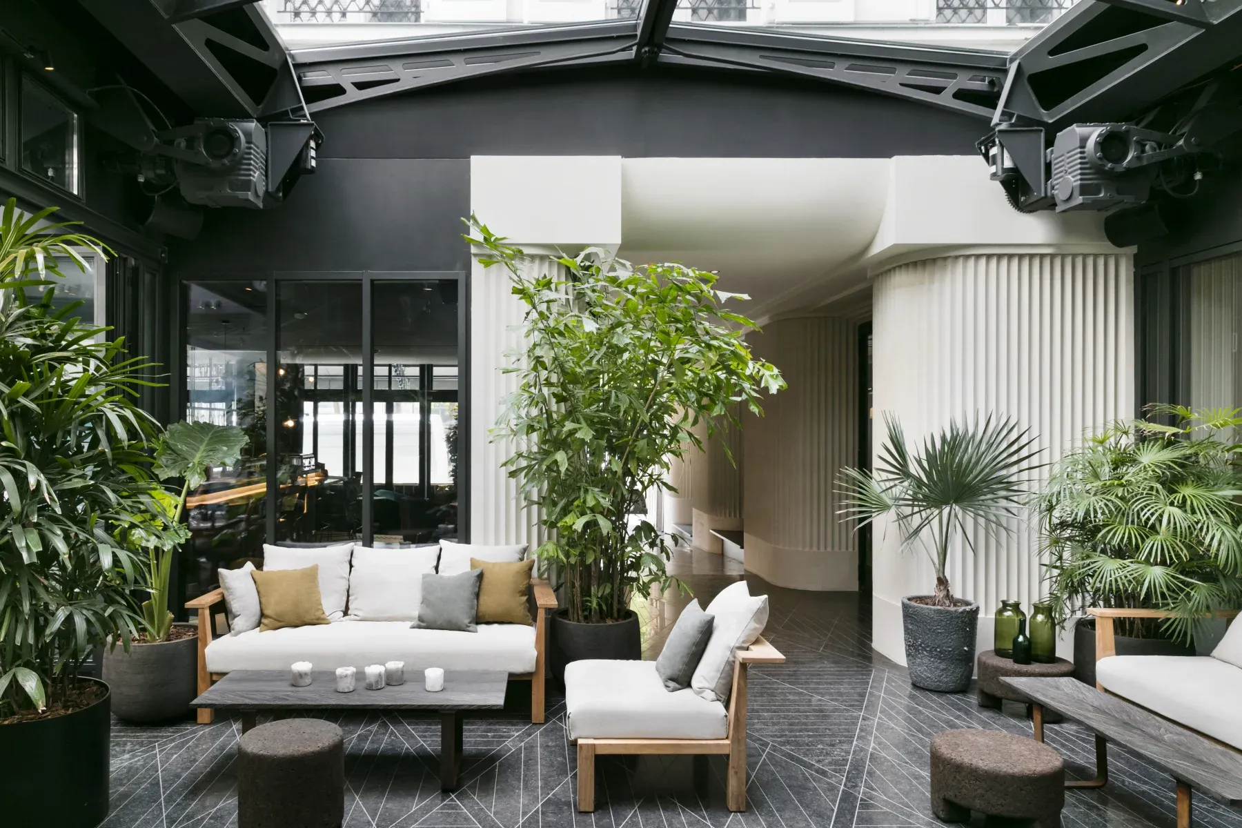 Hôtel National Des Arts, Europe — Covered courtyard lounge with natural light, teak seating, potted palms, dark terrazzo flooring, and curved white architectural details.