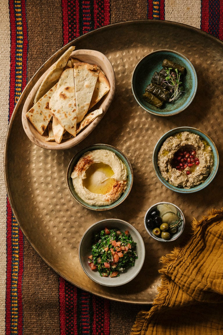 Tama — Hammered brass platter with hummus, baba ganoush, tabbouleh, olives, and warm pita bread on striped textile.