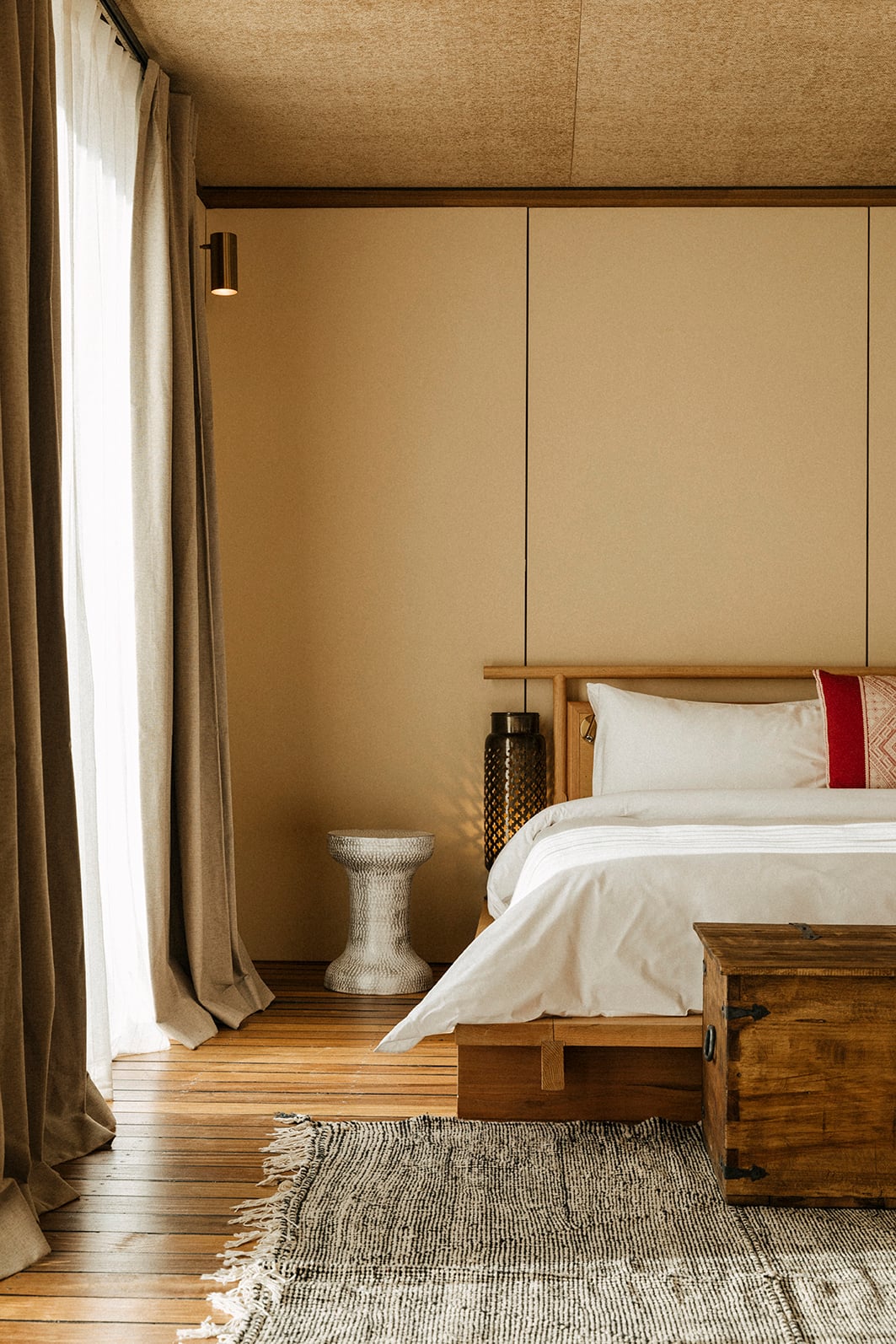 Habitas AlUla — Minimalist bedroom with textured cork ceiling, sage walls, wooden bed frame, cream linens, woven rug, natural light from floor-to-ceiling curtains.