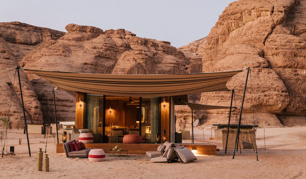 Habitas AlUla, Saudi Arabia — Canvas pavilion with wooden interior nestled between ochre sandstone cliffs, striped awning, ambient lighting, desert landscape.