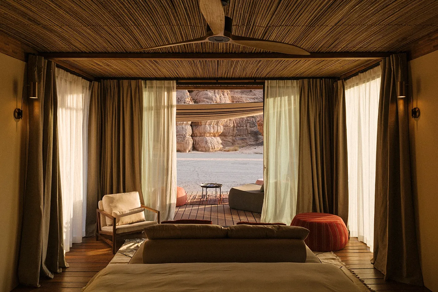 Habitas AlUla — Desert villa interior with wooden ceiling fan, cream linen curtains framing terrace view of sandstone cliffs and wadi landscape, mid-century modern furnishings.
