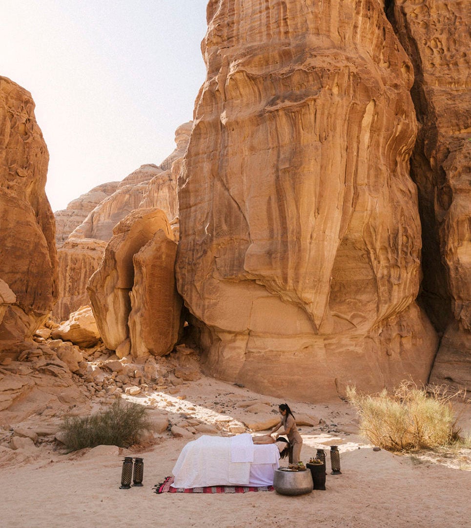 Thuraya Wellness — Woman meditating beneath towering sandstone cliffs in desert landscape, minimalist wellness setup with white fabric and ceramic vessels.