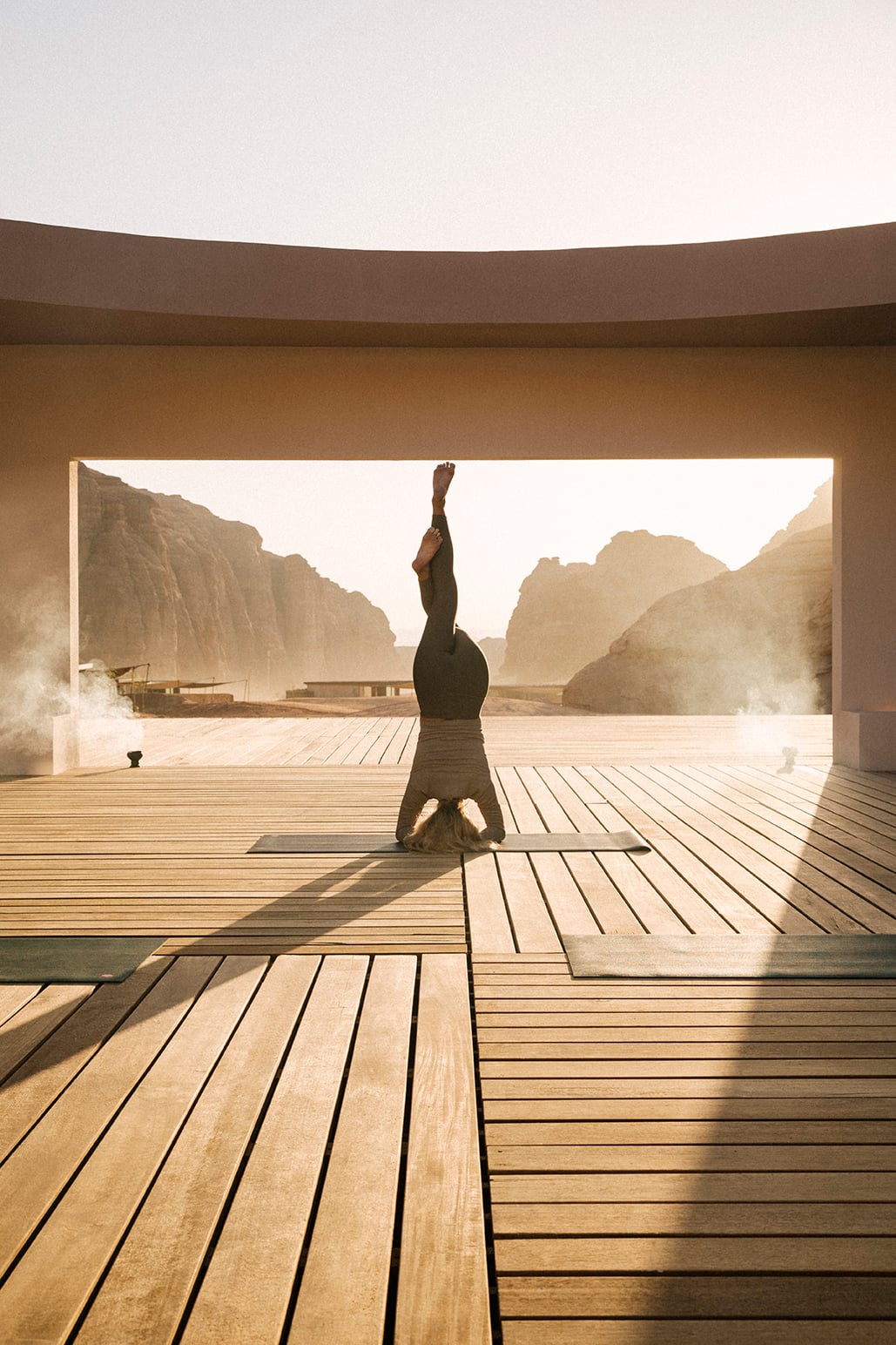 Summer Workshop — Person in headstand on wooden deck overlooking misty limestone cliffs and traditional boats, minimalist architecture framing serene landscape.