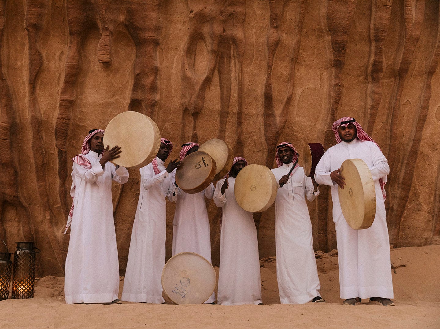 Habitas AlUla, Saudi Arabia — Musicians in white thobes and red ghutra holding traditional frame drums against earthen canyon walls in desert setting.