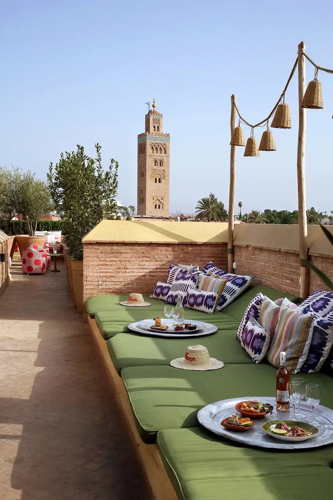 El Fenn Rooftop Bar, Marrakech, Morocco — green cushioned seating with patterned pillows, brass hanging bells, and Koutoubia Mosque minaret view under clear sky.
