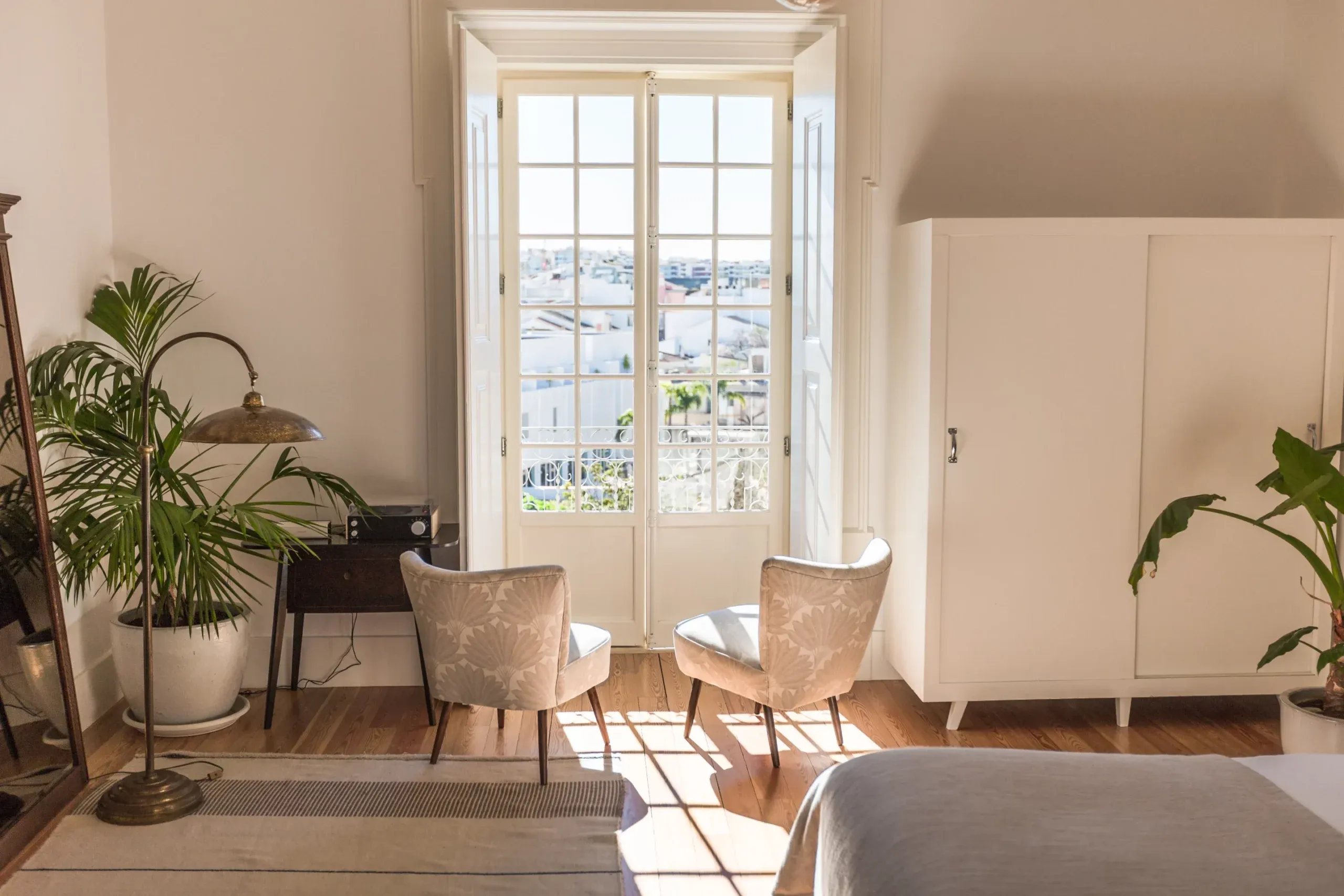 Casa Mãe, Europe — Sunlit bedroom with french doors overlooking cityscape, mid-century chairs, potted plants, natural wood floors, minimalist modern aesthetic.