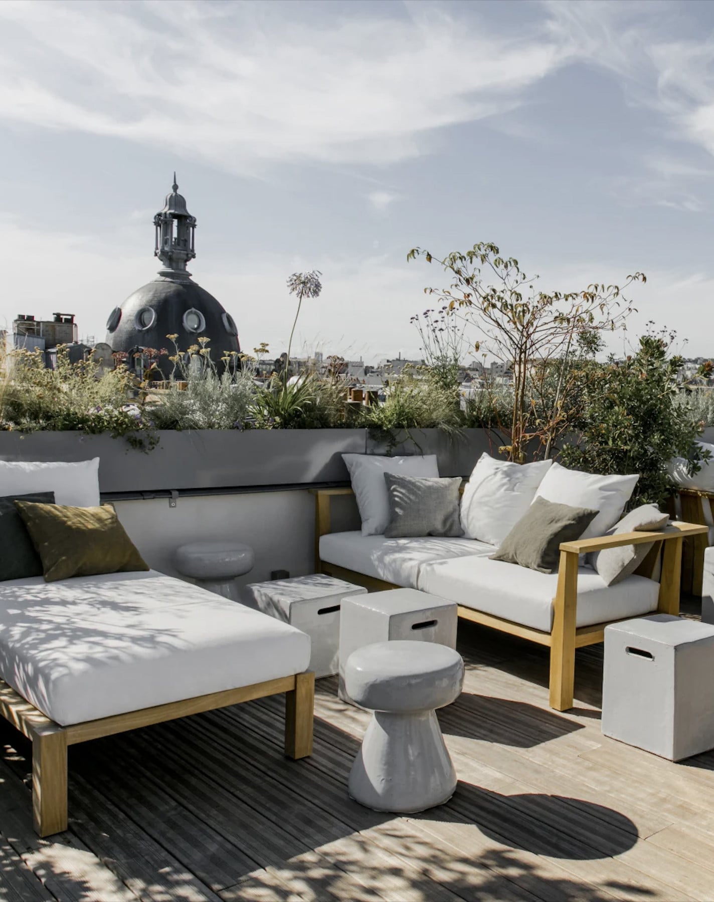 Hôtel National Des Arts, Europe — Rooftop lounge with teak furniture, white cushions, planted containers, and domed architecture beyond, daylit urban vista.