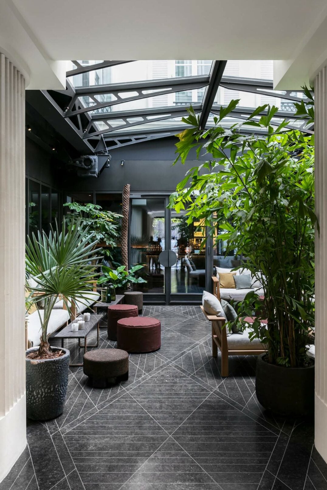 Hôtel National des Arts — Modern covered courtyard with geometric stone flooring, lush green plants, burgundy ottomans, and steel-framed skylight above dark walls.