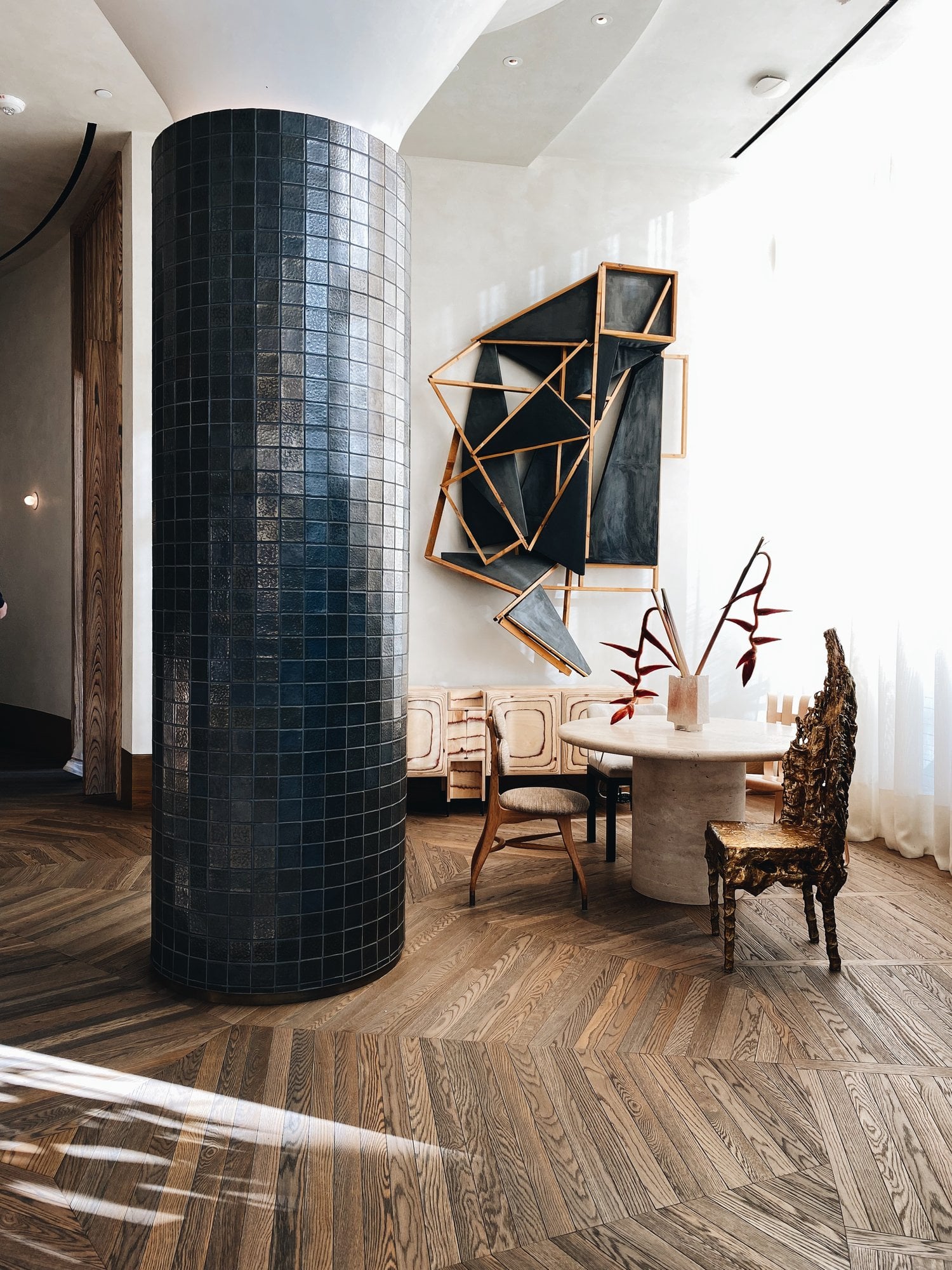 Santa Monica Proper, Los Angeles, USA — Cylindrical dark tile column, wooden herringbone flooring, sculptural wall art with geometric frames, round travertine table, modernist furniture in bright gallery space.