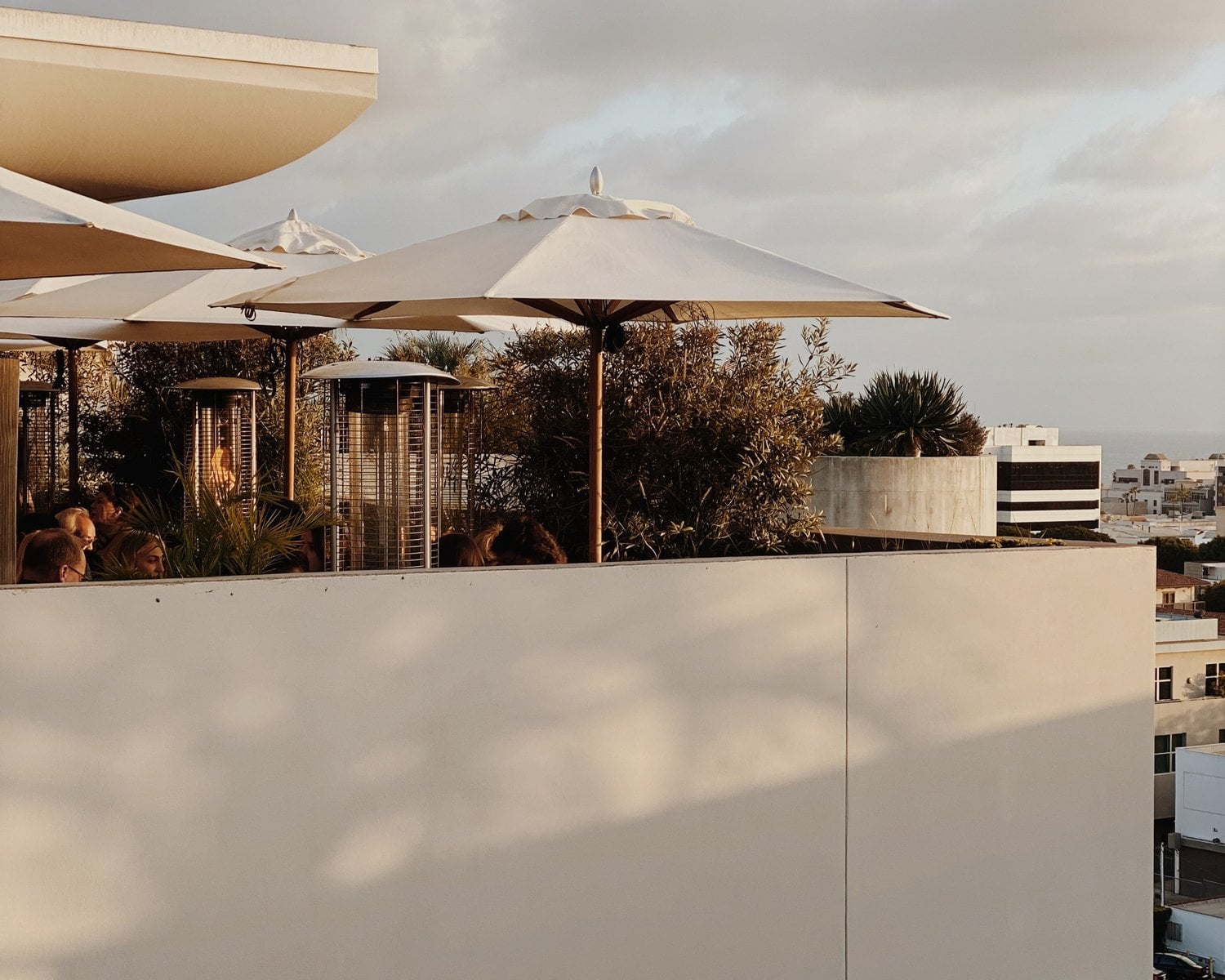 Santa Monica Proper, Los Angeles, USA — Rooftop terrace with cream umbrellas, architectural concrete parapet, olive trees, and city skyline views at dusk.