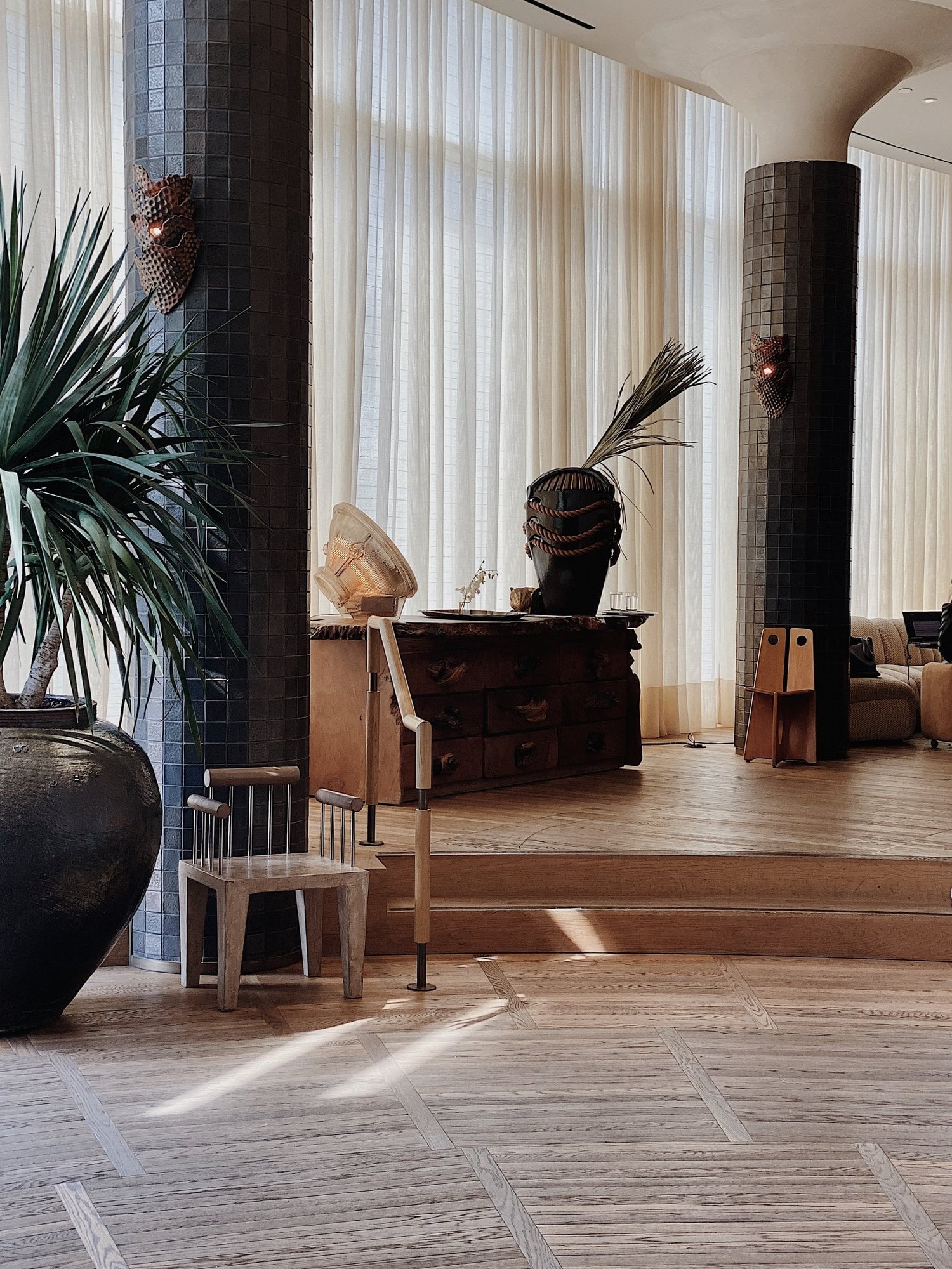 Santa Monica Proper, North America — Minimalist lobby with dark tile columns, natural light through sheer vertical blinds, wooden herringbone flooring, sculptural vessels and potted plants.