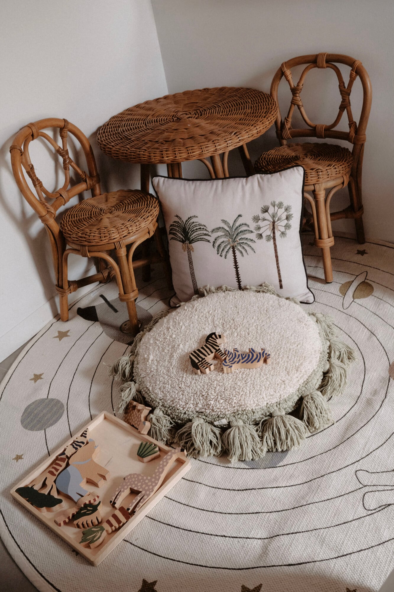 Vista Verde, Balearic Islands — Woven rattan furniture and round cream cushion with tassel trim arranged on patterned floor with palm-print pillow and wooden toy safari set.