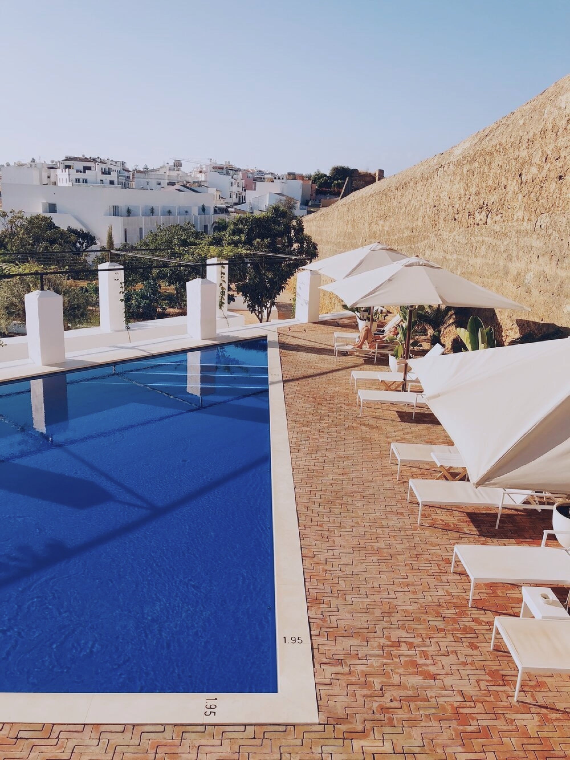 Casa Mãe, Europe — Modernist poolside terrace with cobalt water, whitewashed umbrellas, terracotta herringbone deck, limestone fortification wall, Mediterranean architecture.