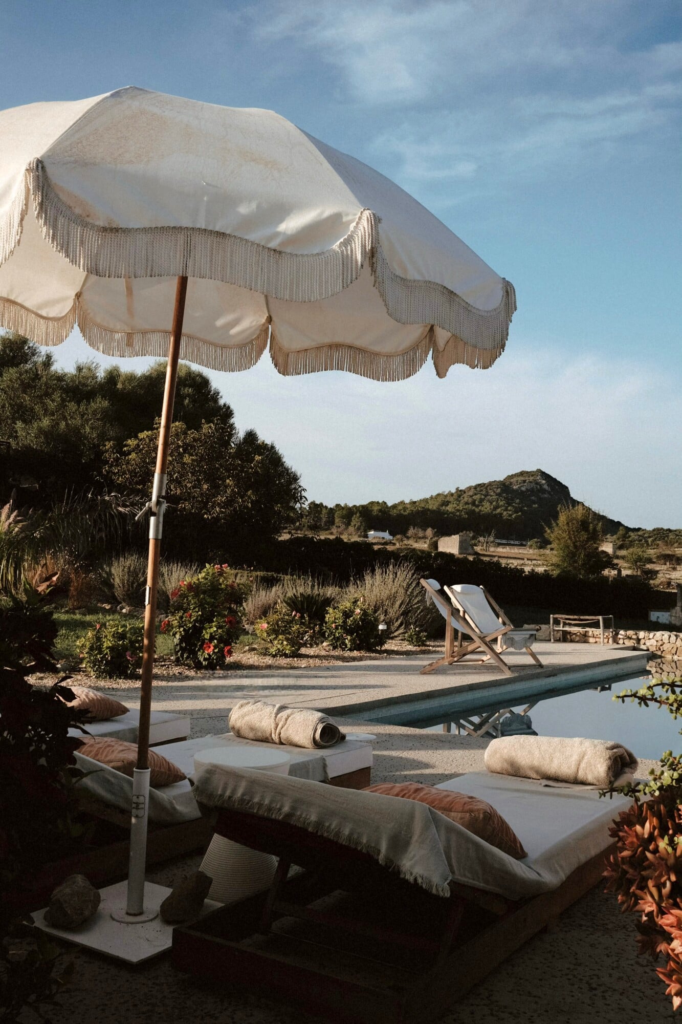 Vista Verde, Balearic Islands — Cream linen parasol with fringe trim casting shadows over lounge beds beside pool, Mediterranean landscape and hills beyond.