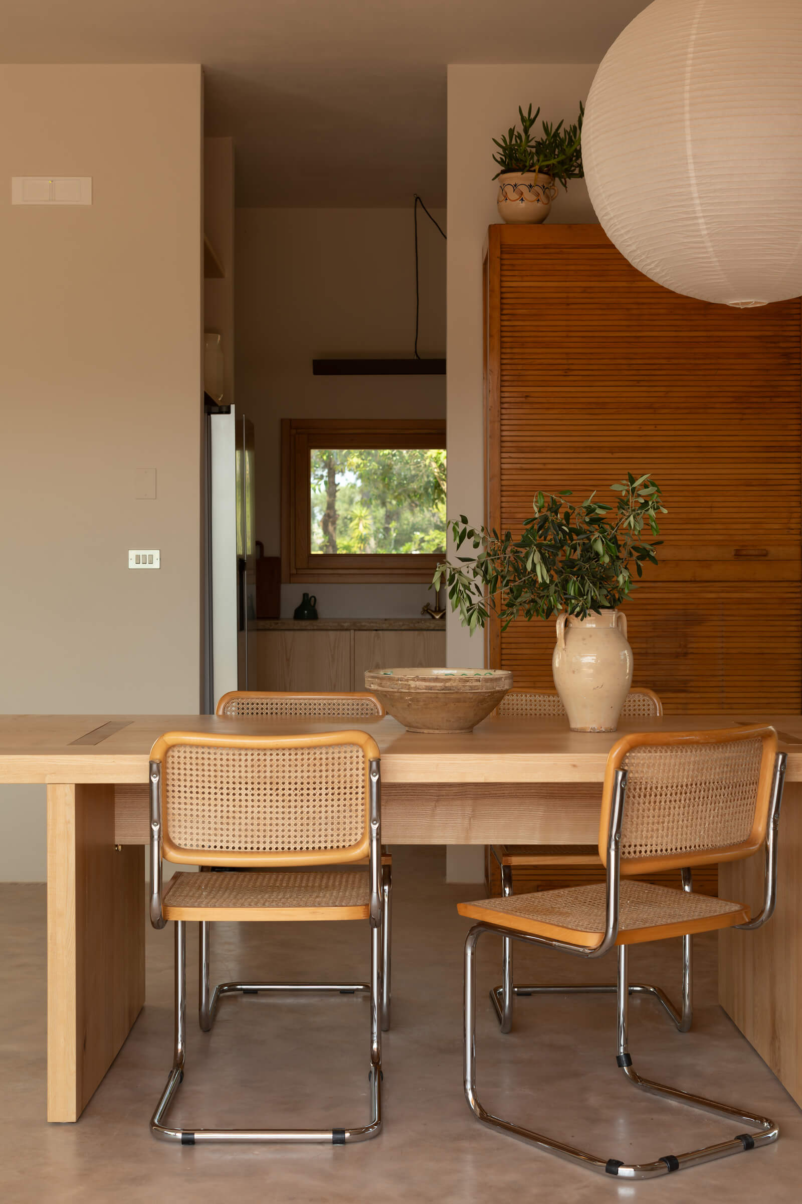 Casanguilla, Europe — Minimalist dining room with light wood table, caned chairs, potted plants, and pendant lamp overlooking garden through window.