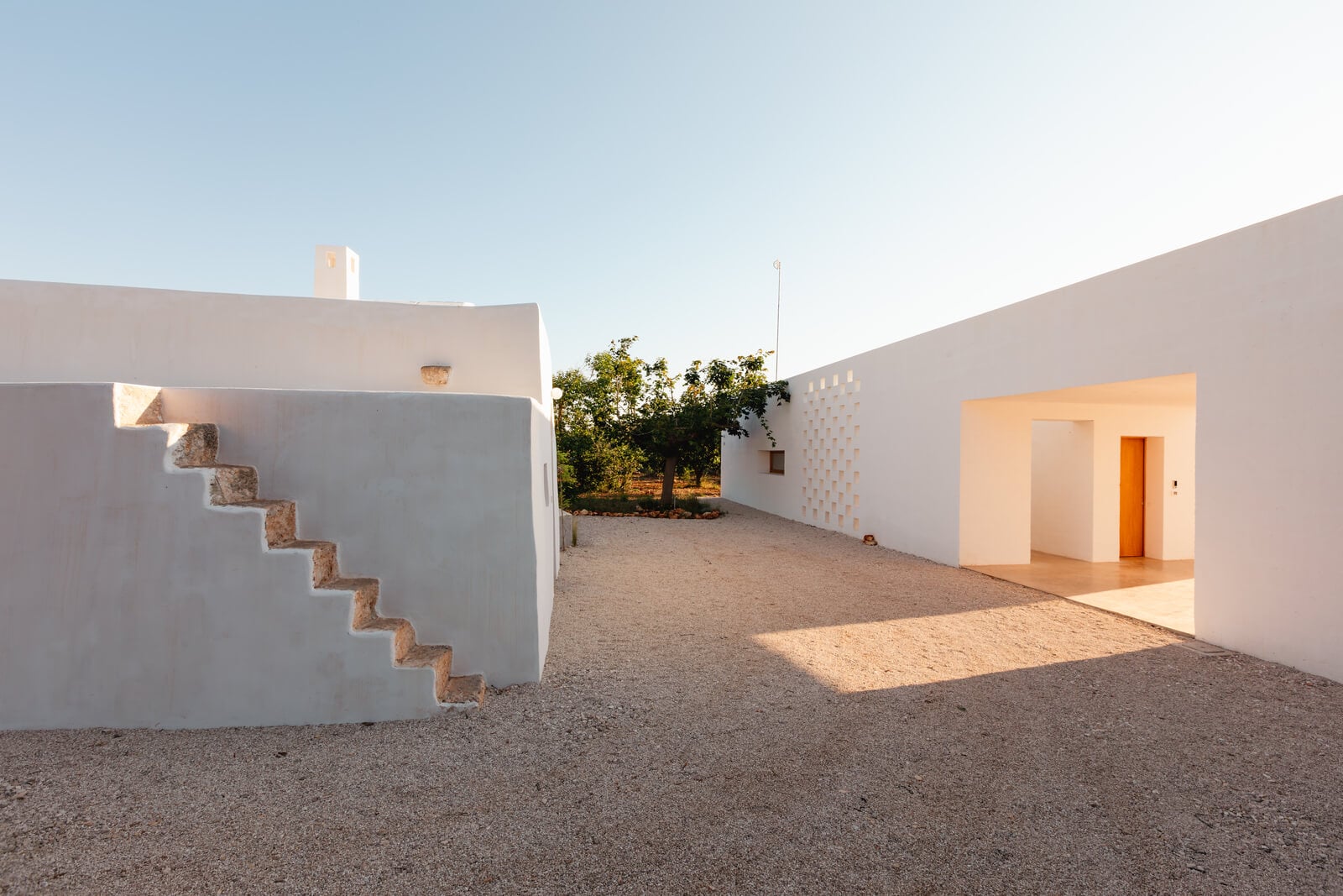 Casanguilla, Europe — Minimalist white courtyard with geometric brick steps, warm golden light, ochre door, and terraced gardens beyond.