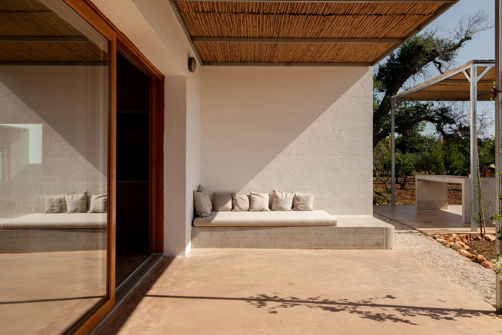 Casanguilla, Europe — Minimalist courtyard with whitewashed walls, wooden pergola, built-in daybed with linen cushions, and dappled sunlight casting shadows on terracotta flooring.