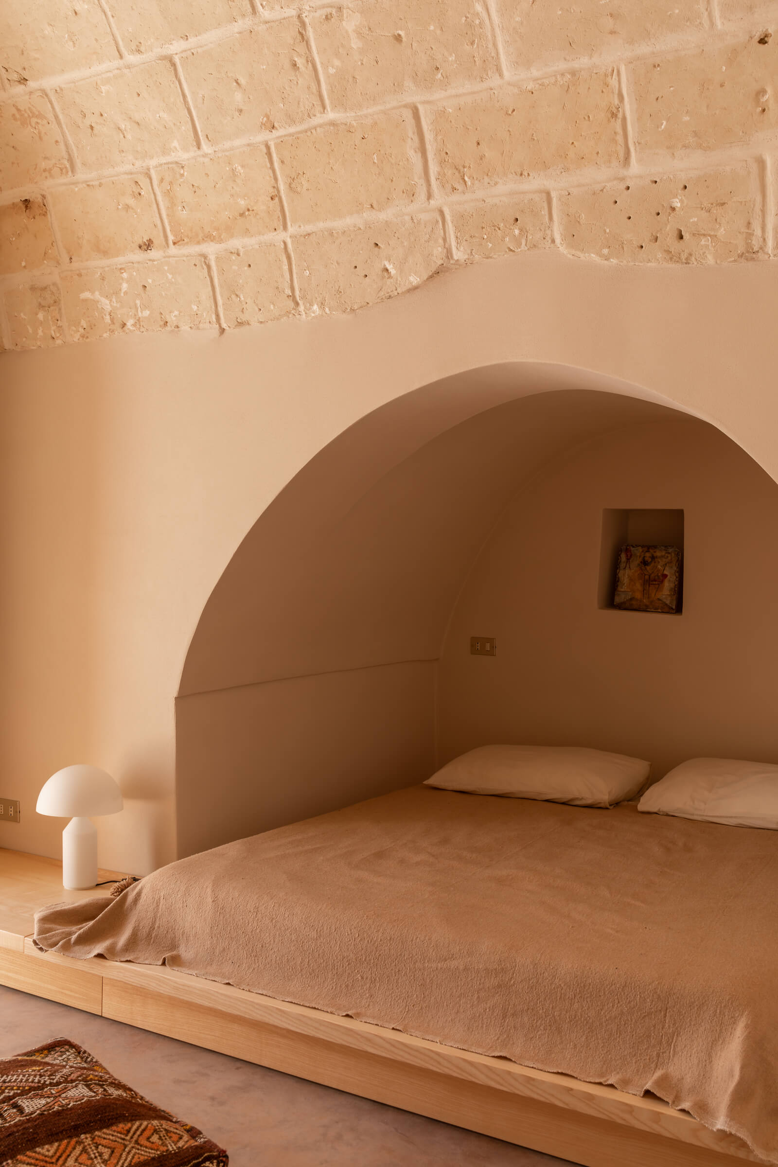 Casanguilla, Europe — Minimalist bedroom with curved plaster archway, terracotta brick ceiling, natural linen bedding, white mushroom lamp, and small window niche in warm neutral tones.